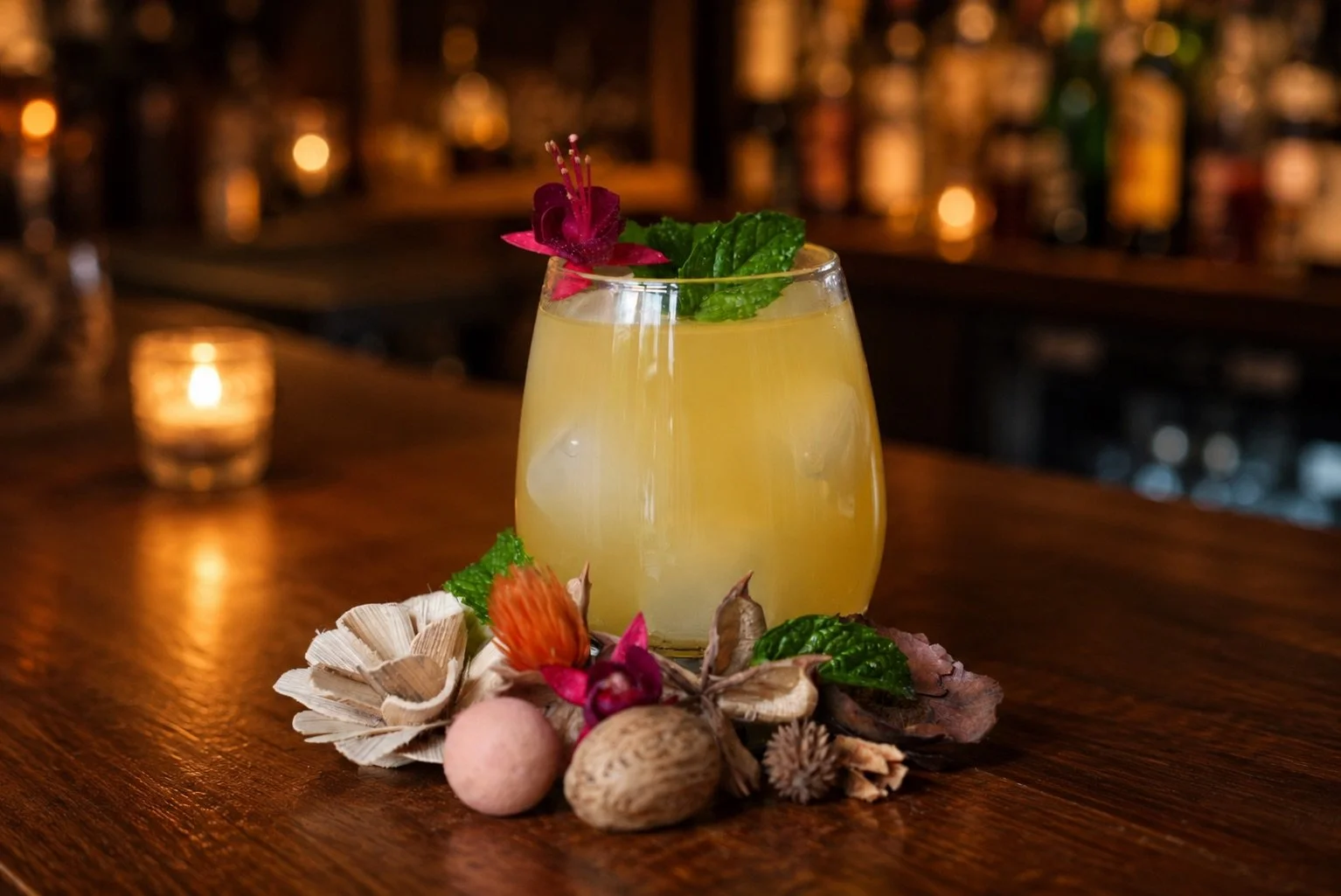 A yellow cocktail garnished with a pink flower and mint leaves, surrounded by decorative natural elements such as dried flowers, seed pods, and nuts on a wooden bar counter. The background features a softly lit bar scene.
