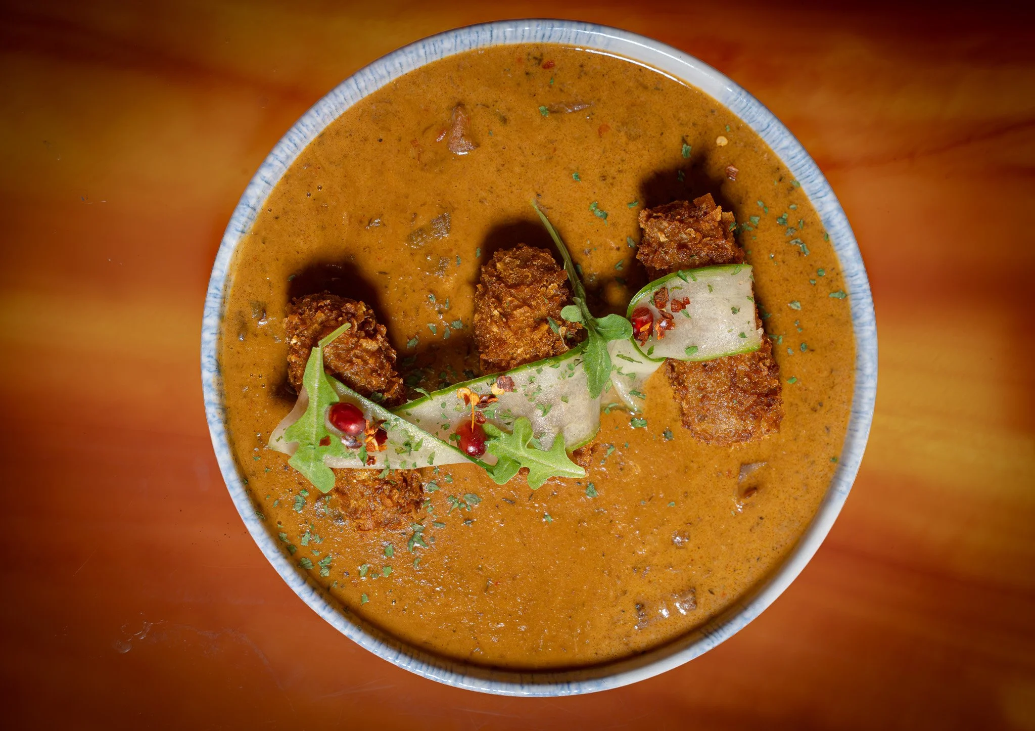 A bowl of Indian curry garnished with green herbs and red chili flakes, topped with fried fritters and a slice of thin cucumber.