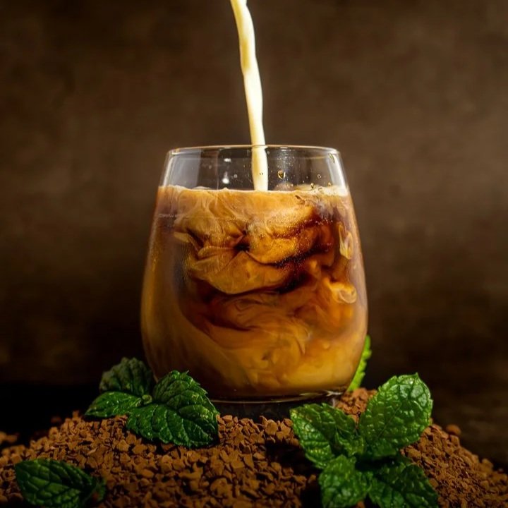 Pouring cream into a glass of iced coffee with ice cubes, garnished with mint leaves.