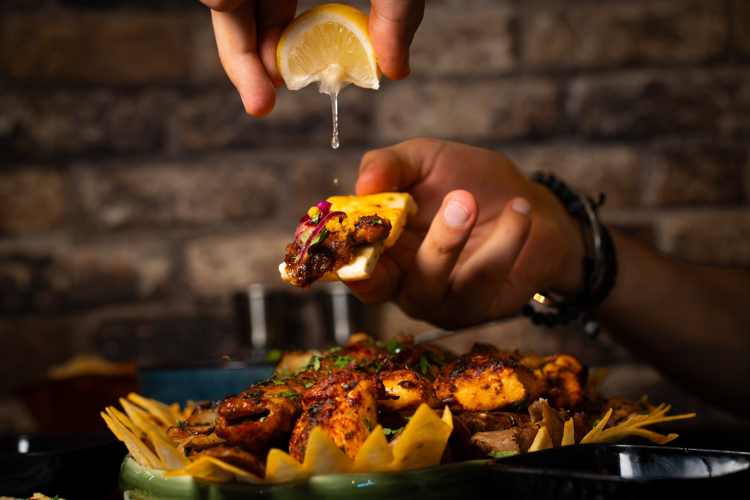 A person squeezing lemon over a plate of chicken tikka with vegetables, garnished with herbs, against a dark background.