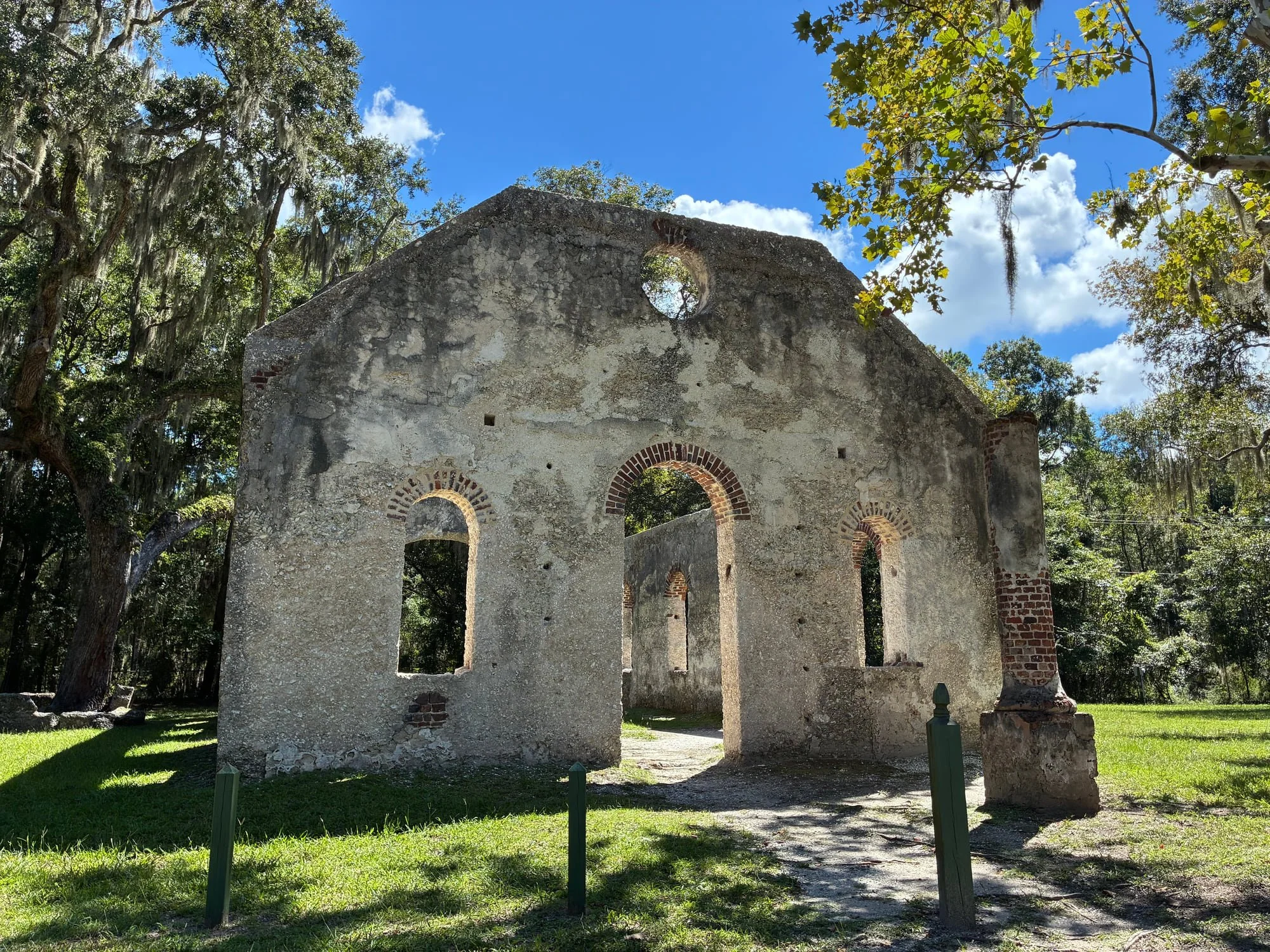 Front view of the roofless Chapel of Ease ruins beneath live oaks and a blue sky