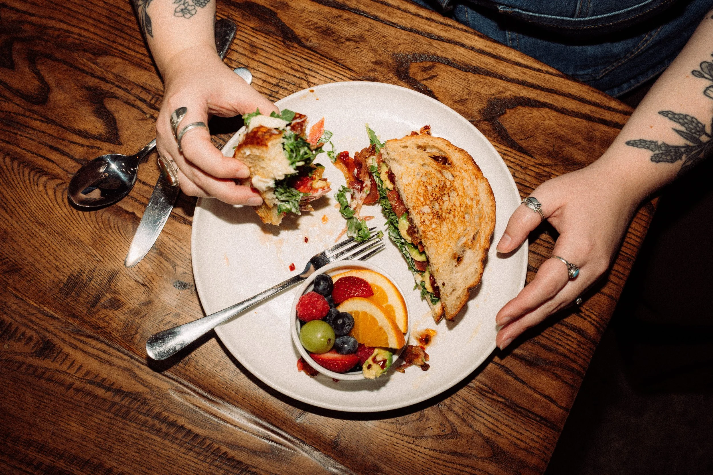 Close-up of a person holding a sandwich with a side of mixed fruits on a white plate, on a wooden table.