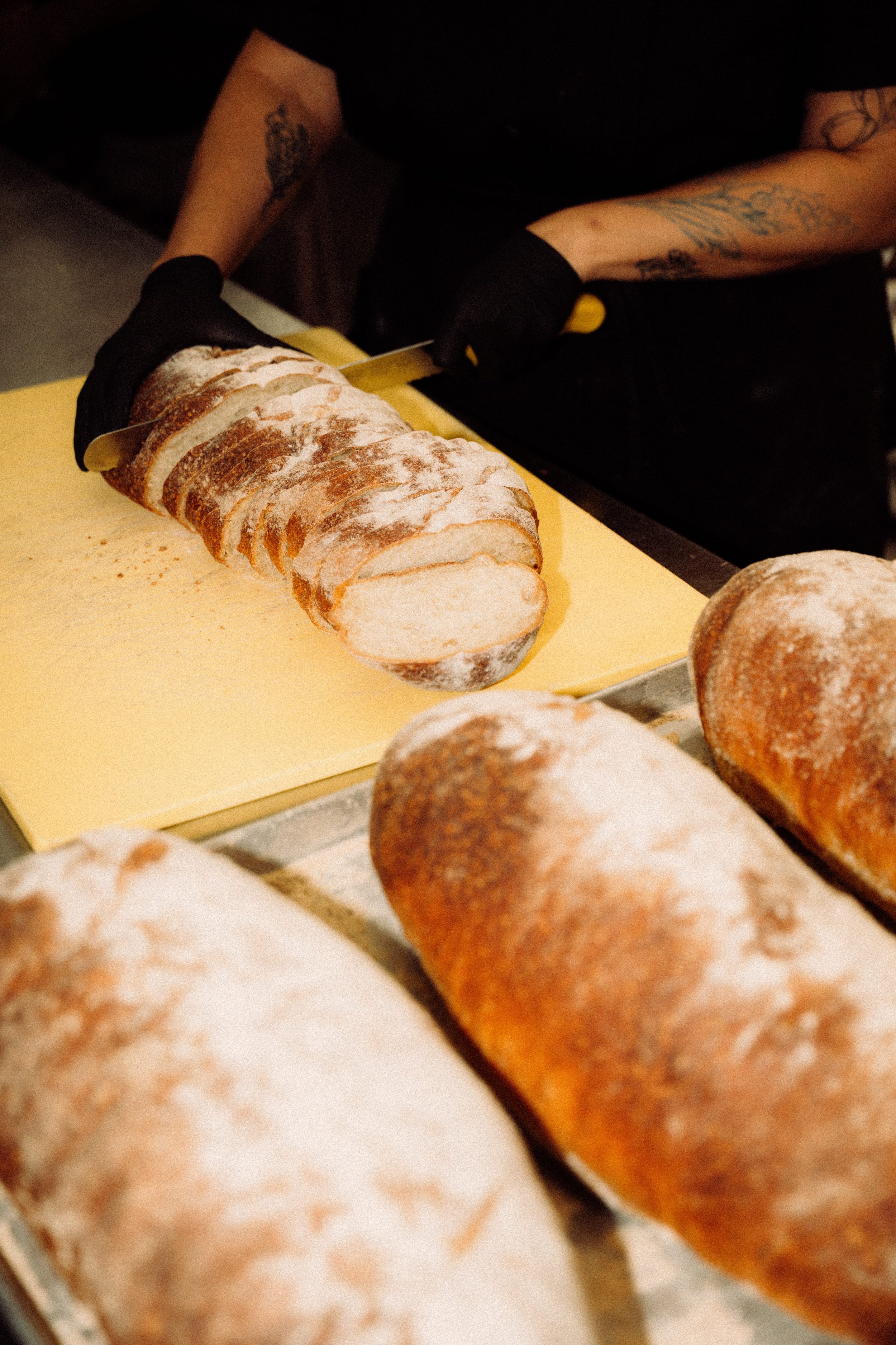 A person slicing a loaf of bread on a yellow cutting board. The person is wearing black gloves and has tattoos on their arms.