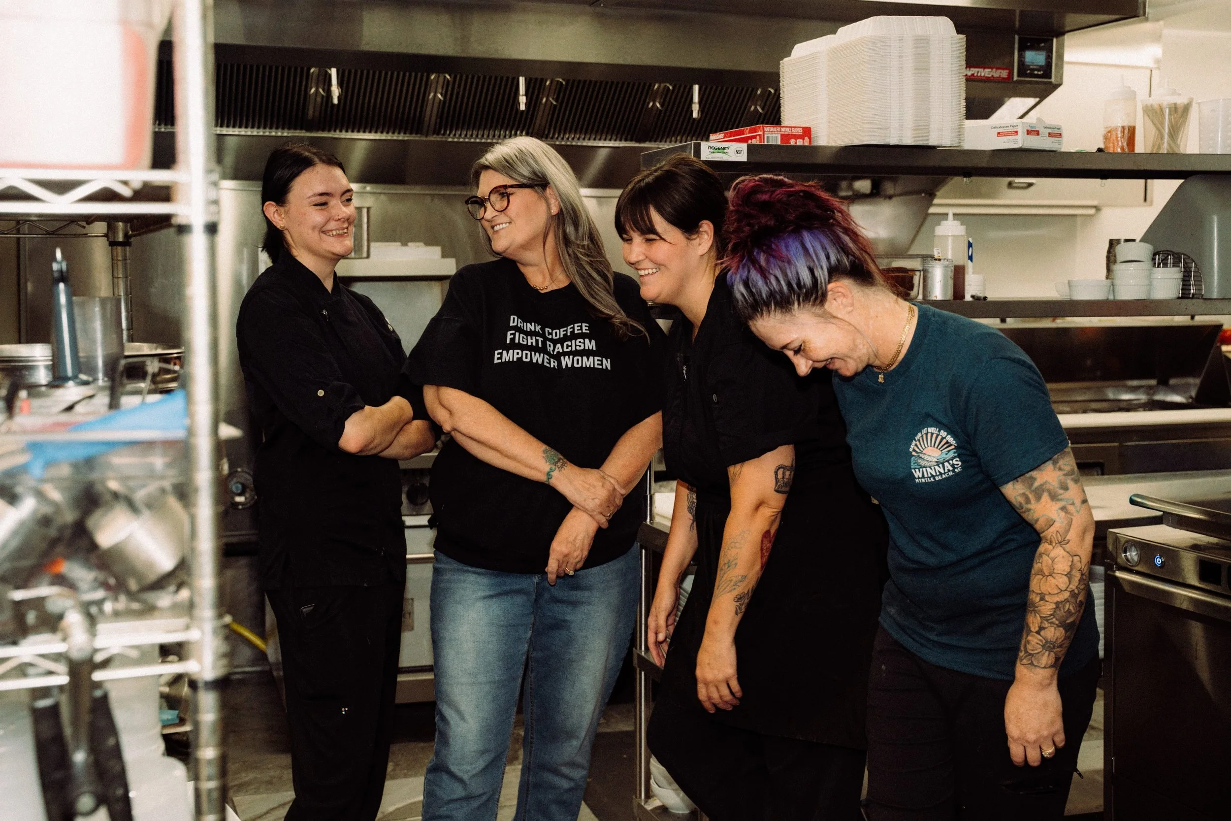 Four women standing in a commercial kitchen, smiling and laughing together.