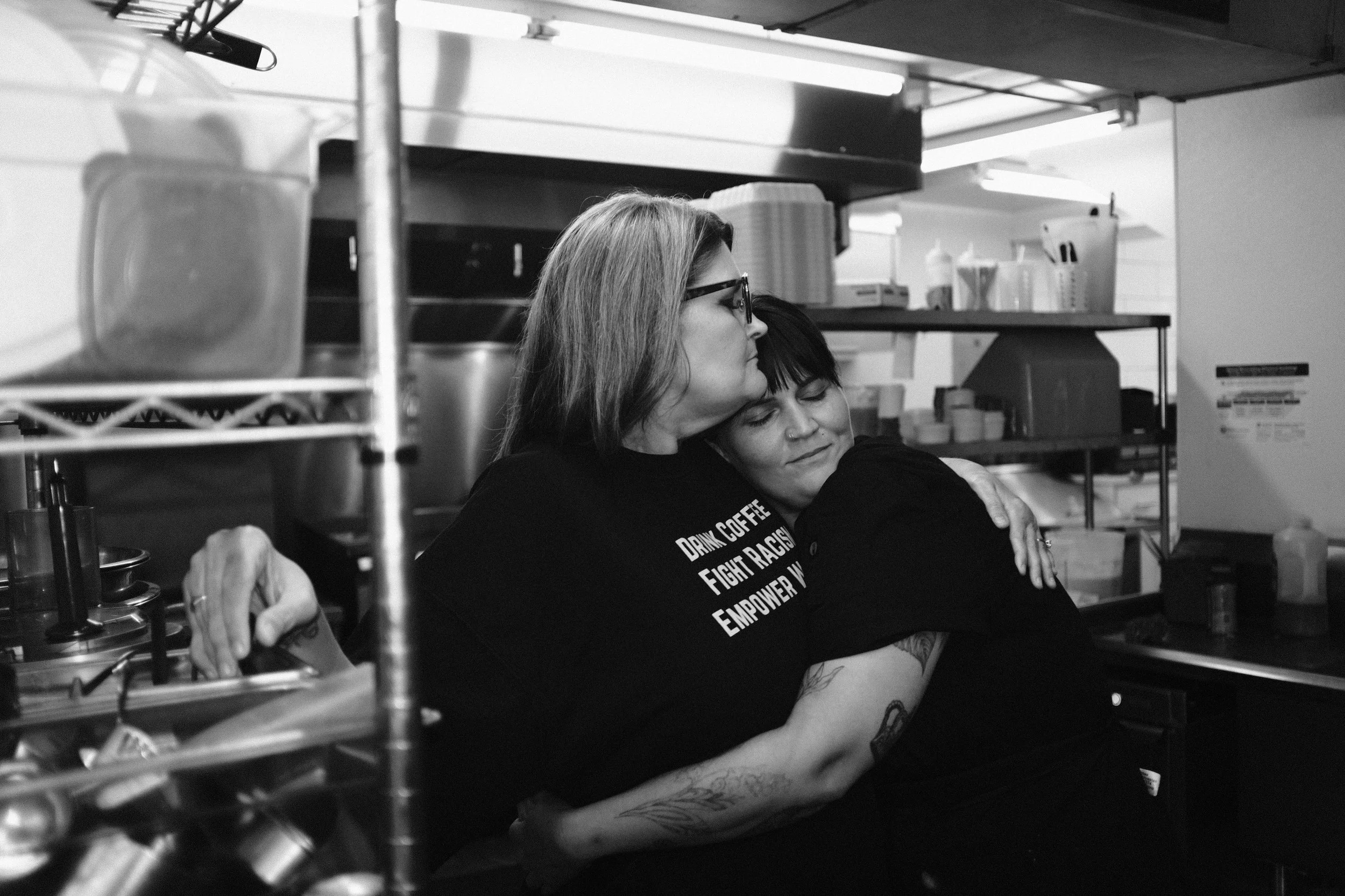 Two women embracing each other in a kitchen, one with glasses and long hair, and another with short dark hair, with kitchen shelves and utensils in the background.