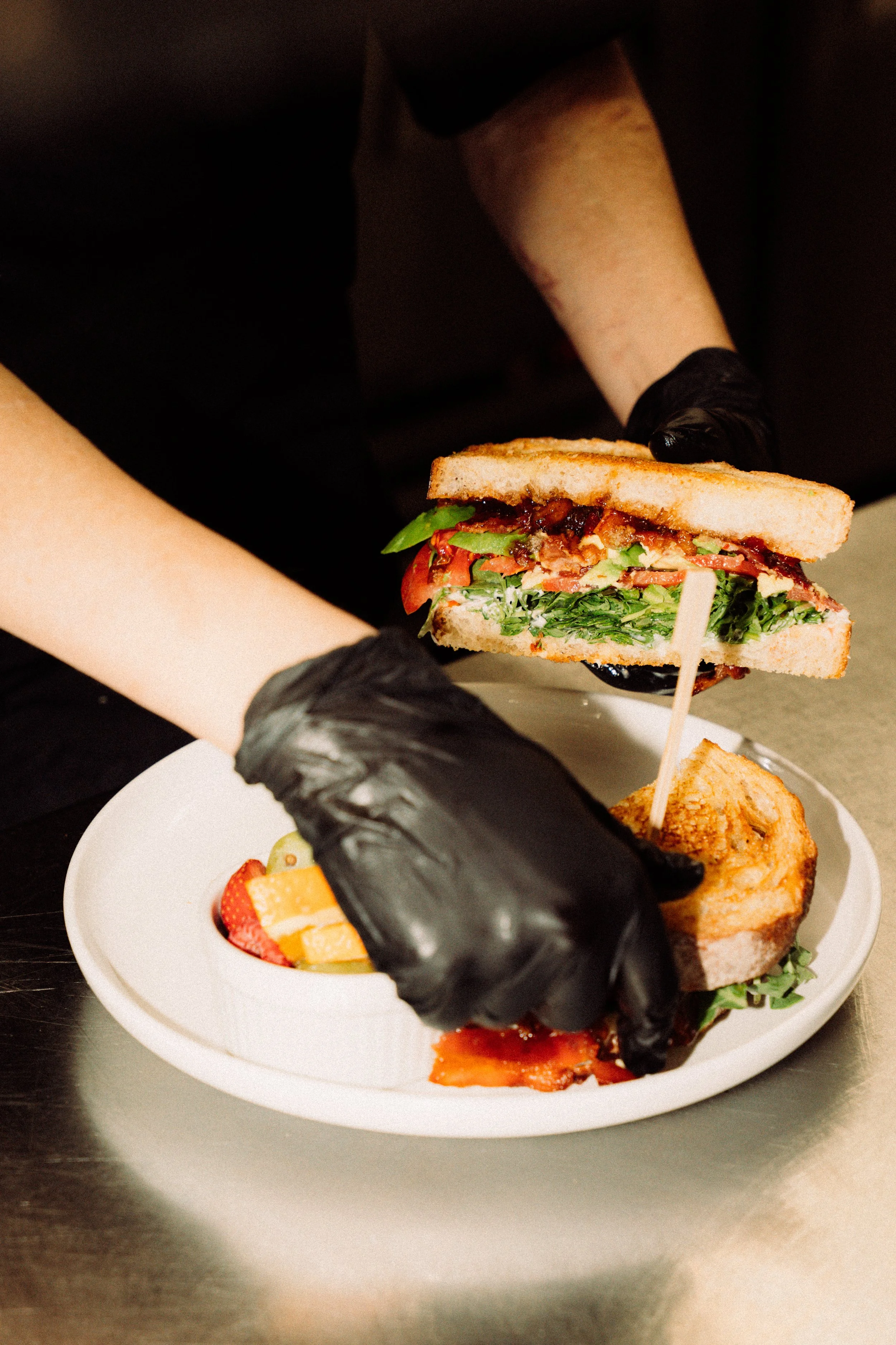 A person holding a sandwich with lettuce, tomato, bacon, and other ingredients, with a black glove on their hand. There is a plate with fruits and possibly a piece of cake or bread in front of them.