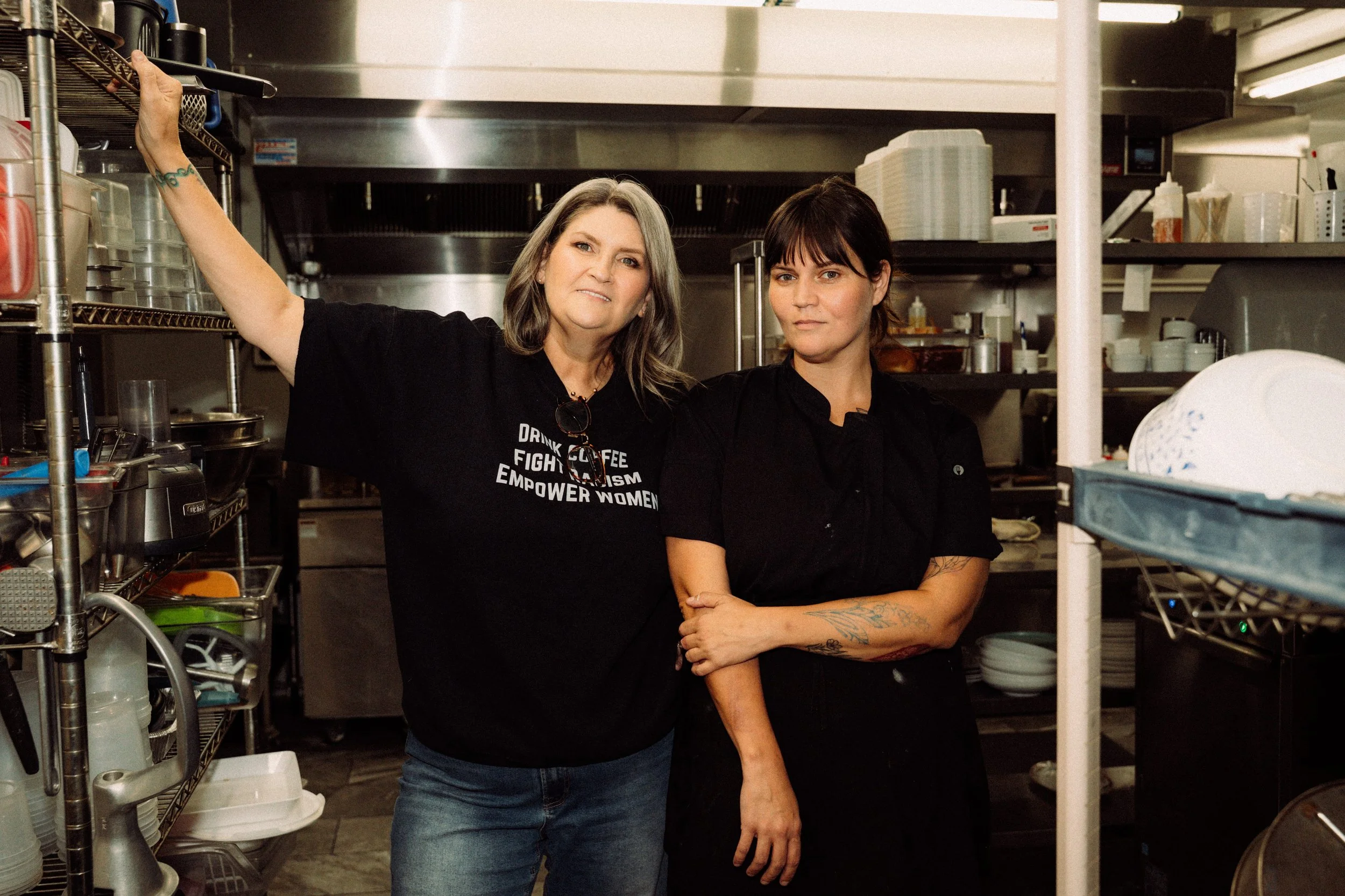 Two women are standing in a kitchen or restaurant environment, with shelves and kitchen equipment around them. One woman has gray hair, is wearing a black T-shirt with text, and is reaching onto a shelf. The other woman has dark hair with bangs, tattoos on her arm, and is standing with her arms crossed, both looking at the camera.
