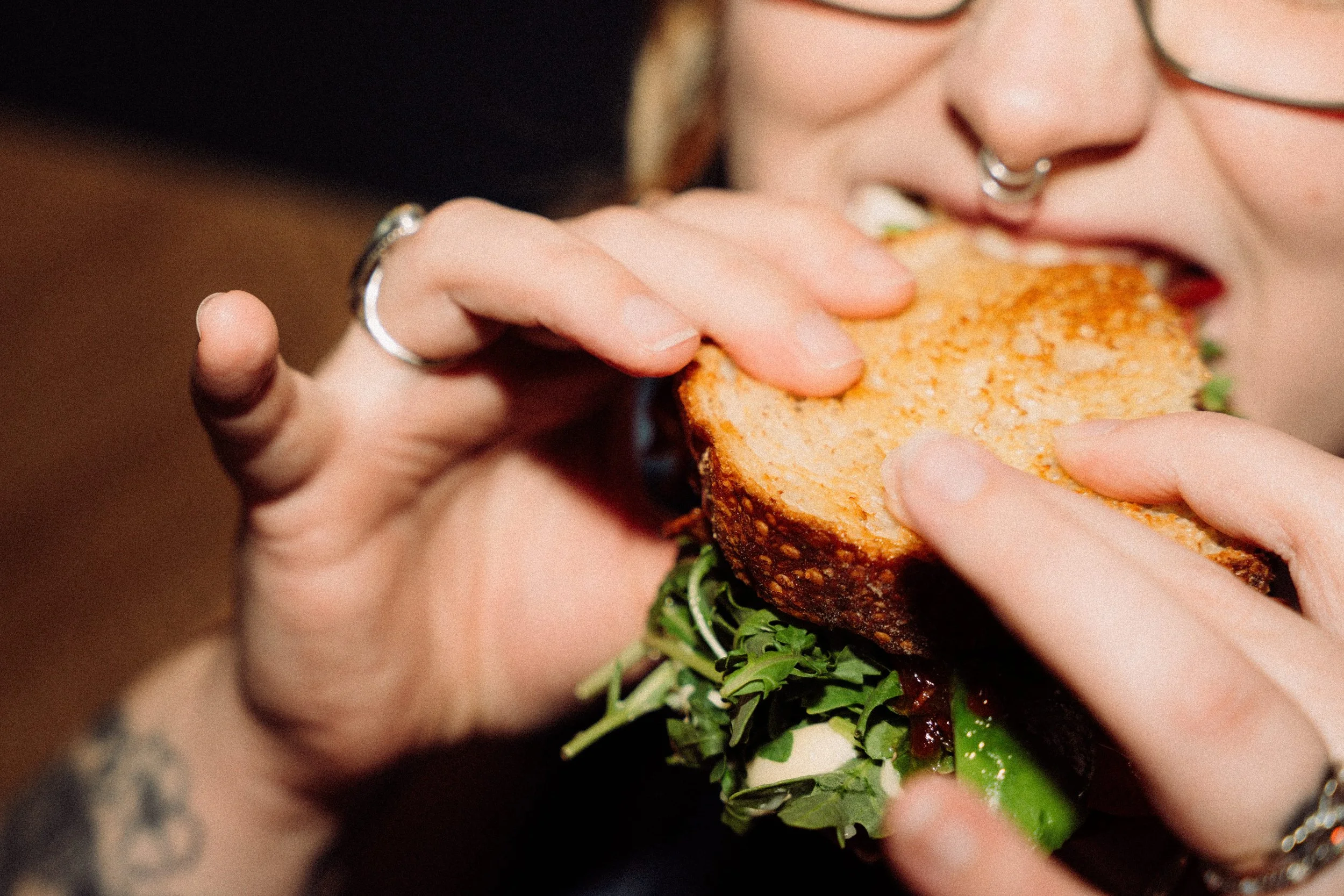 Close-up of a person biting into a sandwich with lettuce, tomato, and a toasted bun, with visible tattoos and rings on their fingers.