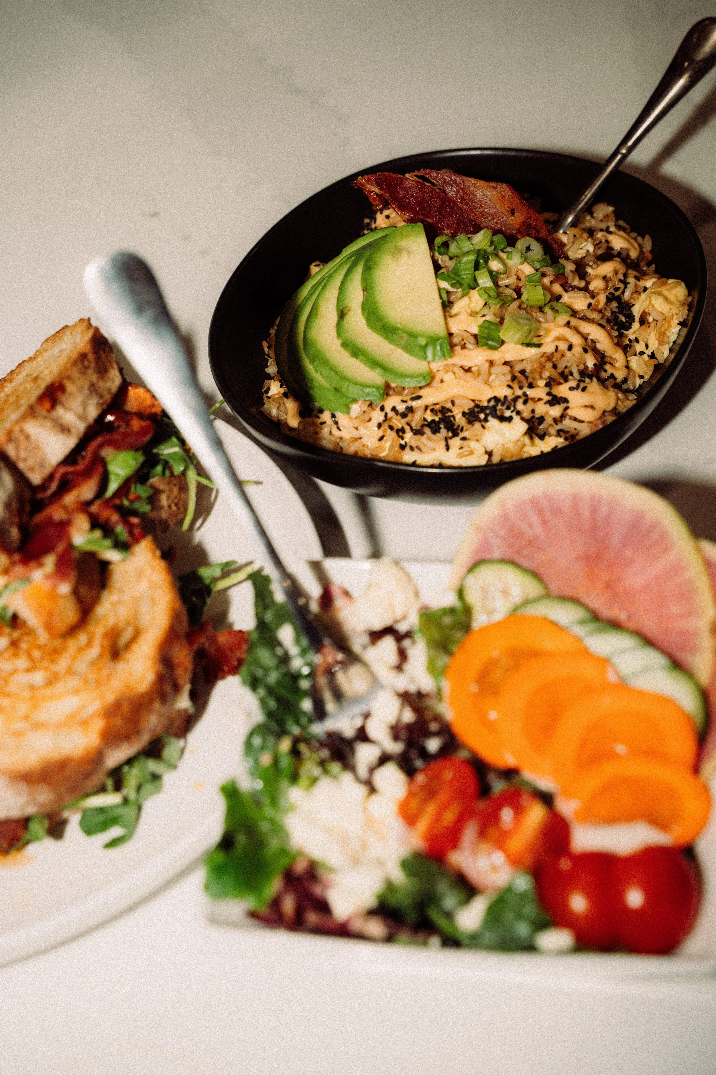 Close-up of a bowl with avocado slices, chopped green onions, bacon, rice, and sesame seeds on top. Part of a plate with sliced fruits and vegetables are visible in the foreground.