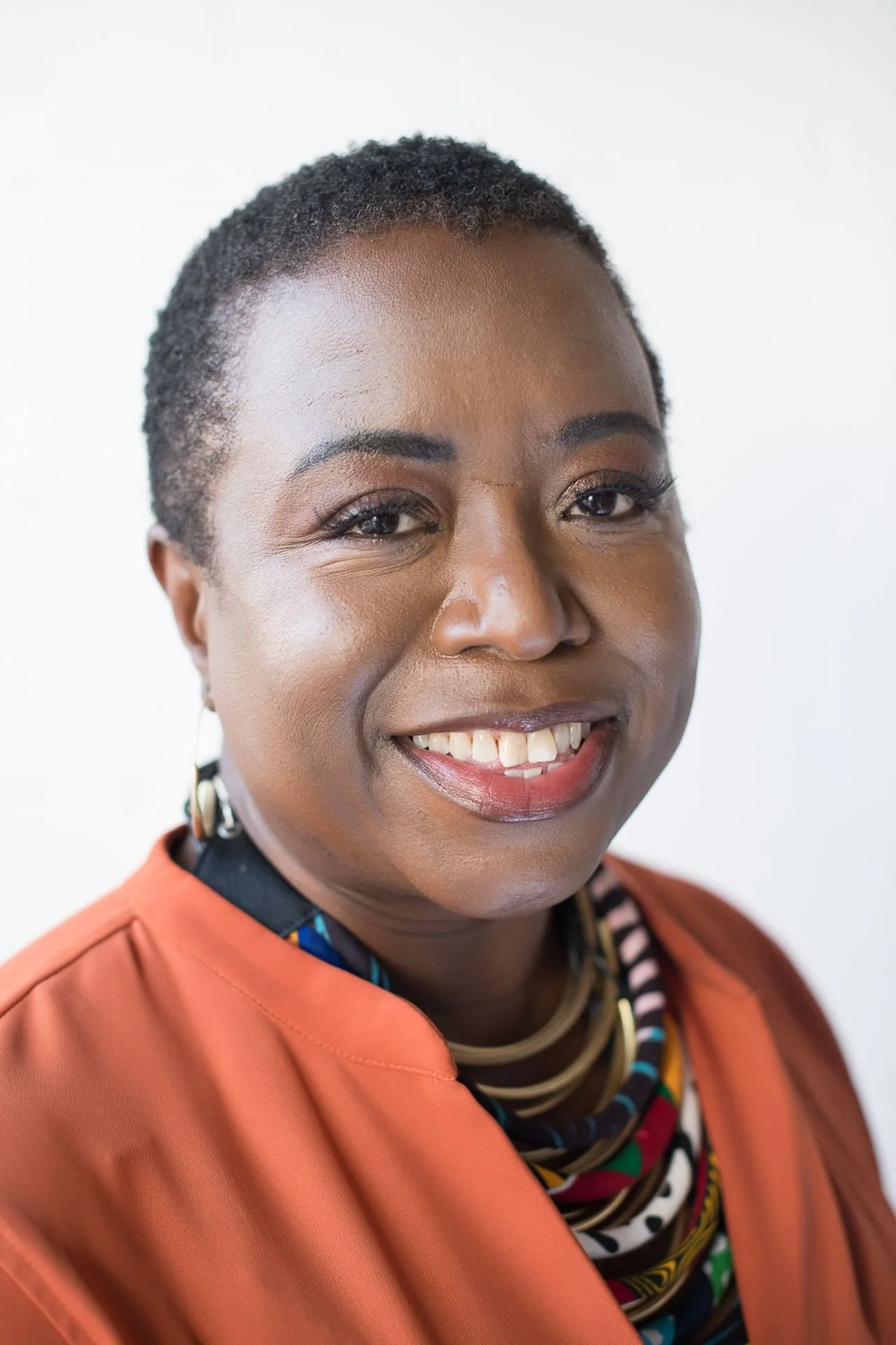 A smiling woman with short curly hair, wearing an orange blazer, gold jewelry, and a multicolored patterned shirt, against a plain white background.