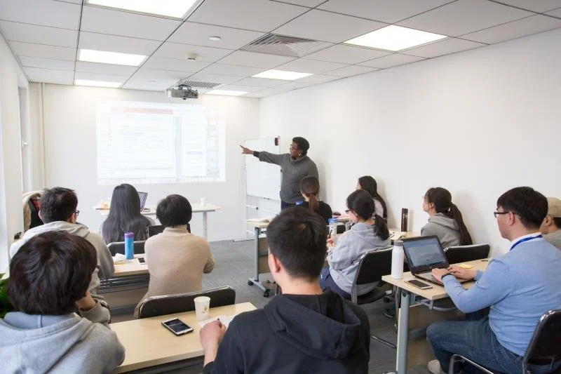 A classroom or conference room with a man giving a presentation to a group of people seated at desks, some using laptops or tablets.