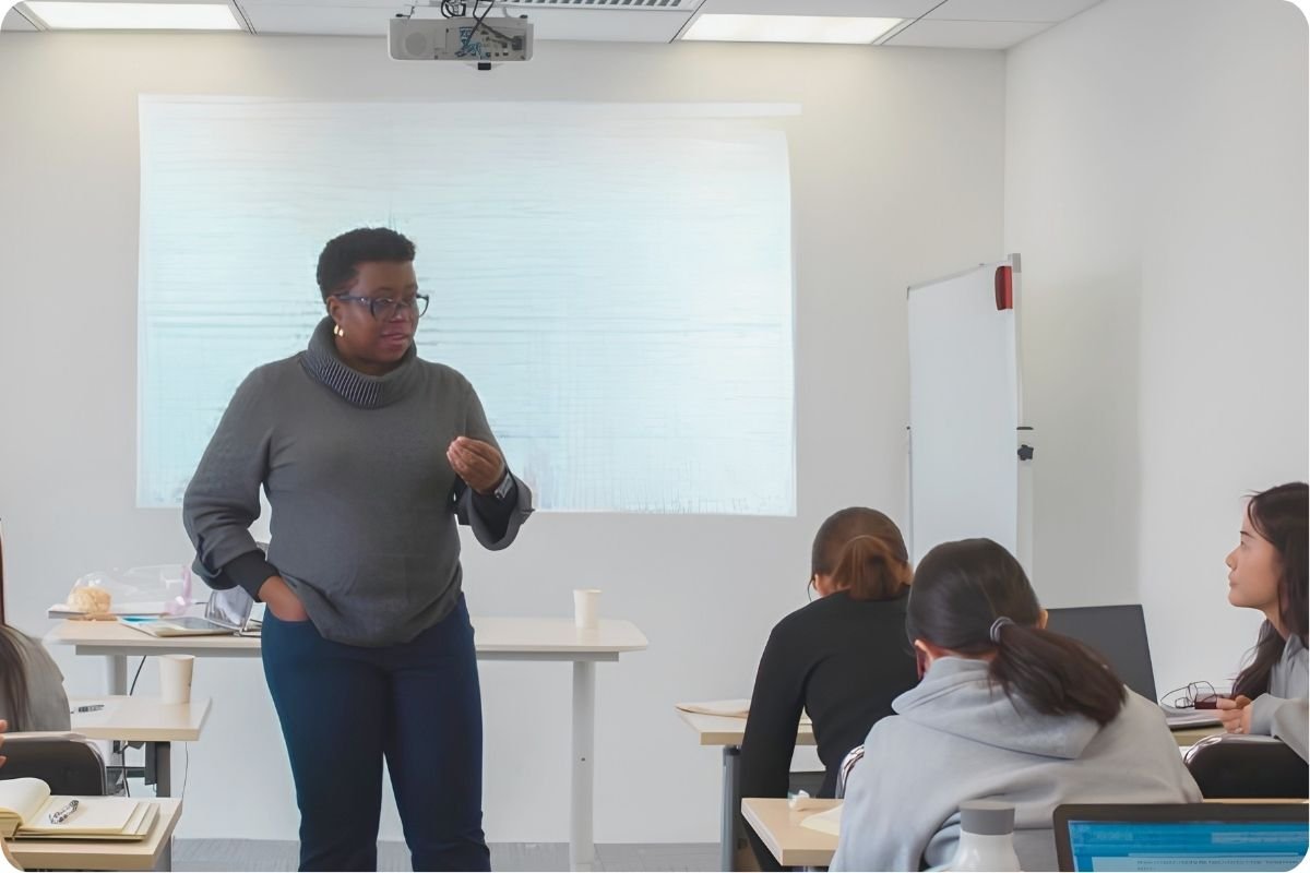 A woman standing in front of a classroom, giving a presentation to students seated at desks, with a whiteboard and projector screen behind her.