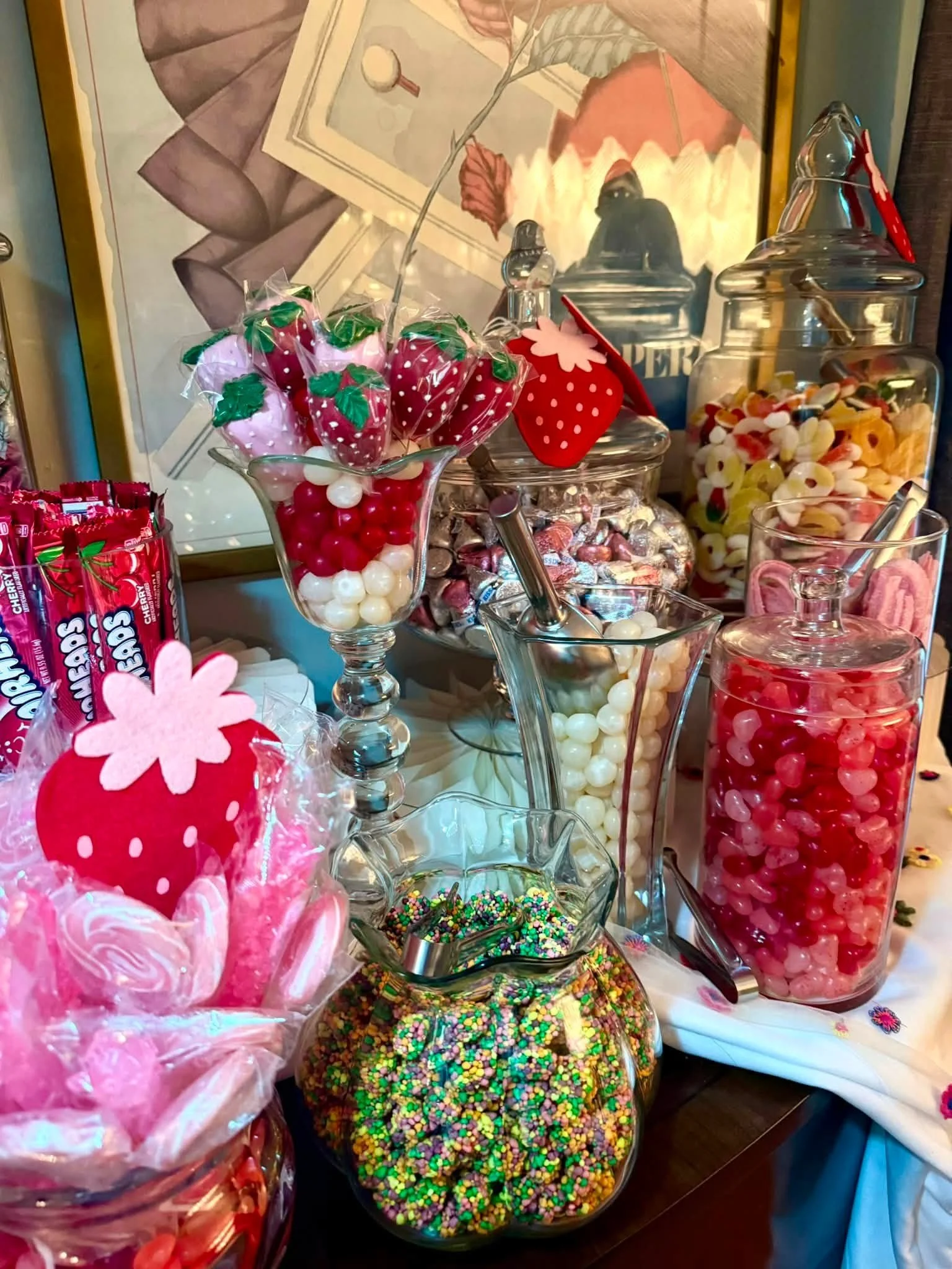 Assorted jars of colorful candies, including gummies and chocolate-covered treats, on a dessert table with a framed artwork in the background.