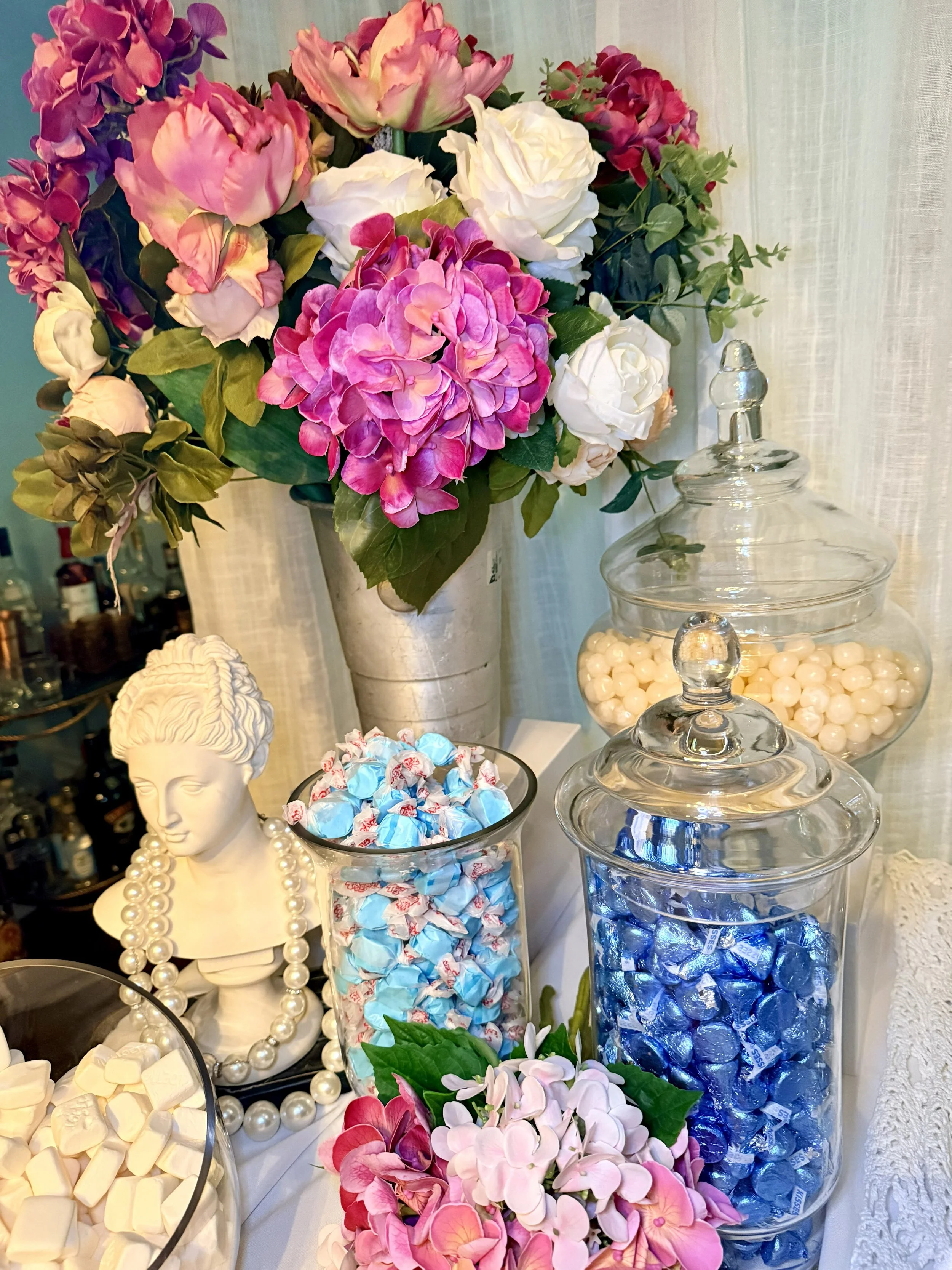 A table display of decorative items including a floral arrangement with pink, purple, white, and beige artificial flowers, a white ceramic bust of a woman with pearl necklaces, and glass jars filled with assorted candies in white, blue, and cream col