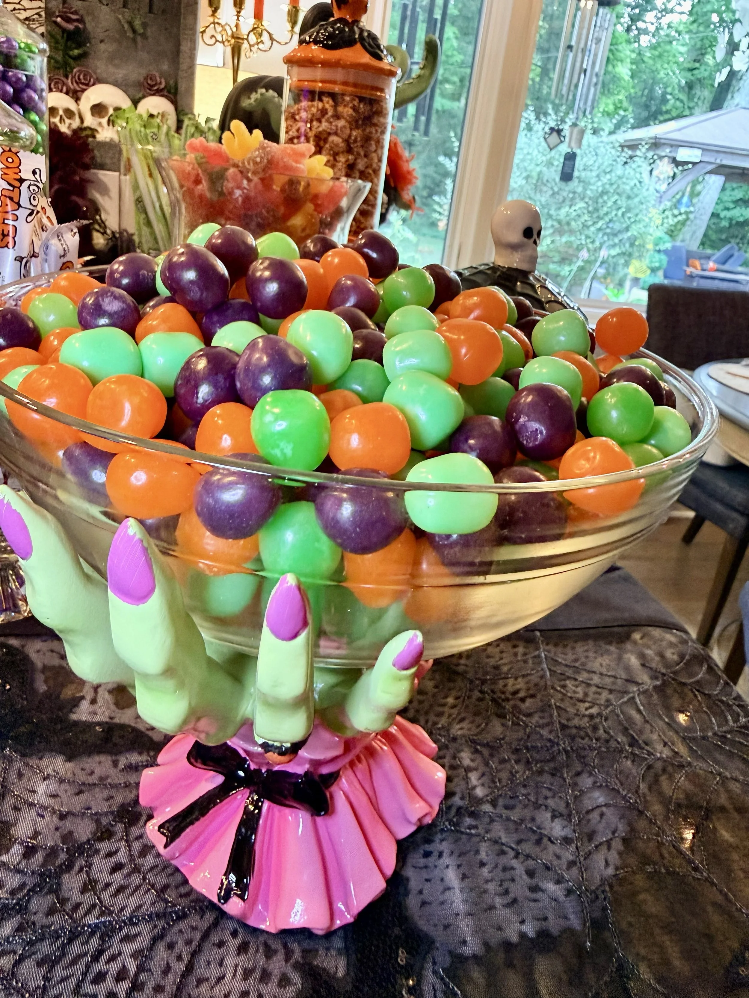Decorative bowl filled with orange, green, and purple artificial grapes, with a skeleton figure in a black cloak beside it on a lace tablecloth, with a Halloween-themed background.