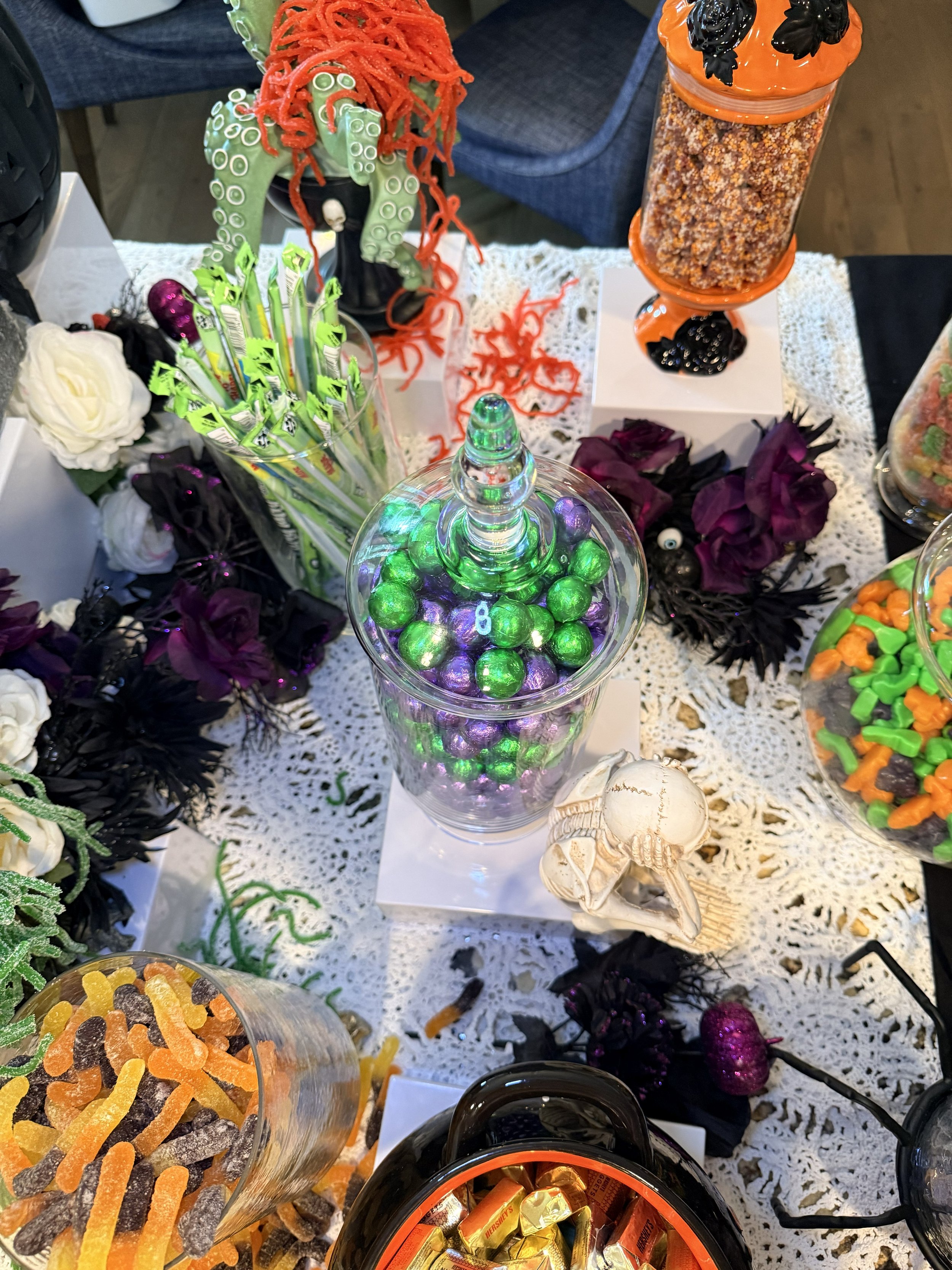 Decorative display of Halloween-themed candy and decorations, including a jar of mixed chocolates, a bowl of gummies, a skeleton figurine, and various Halloween-themed floral arrangements, all arranged on a lace tablecloth.