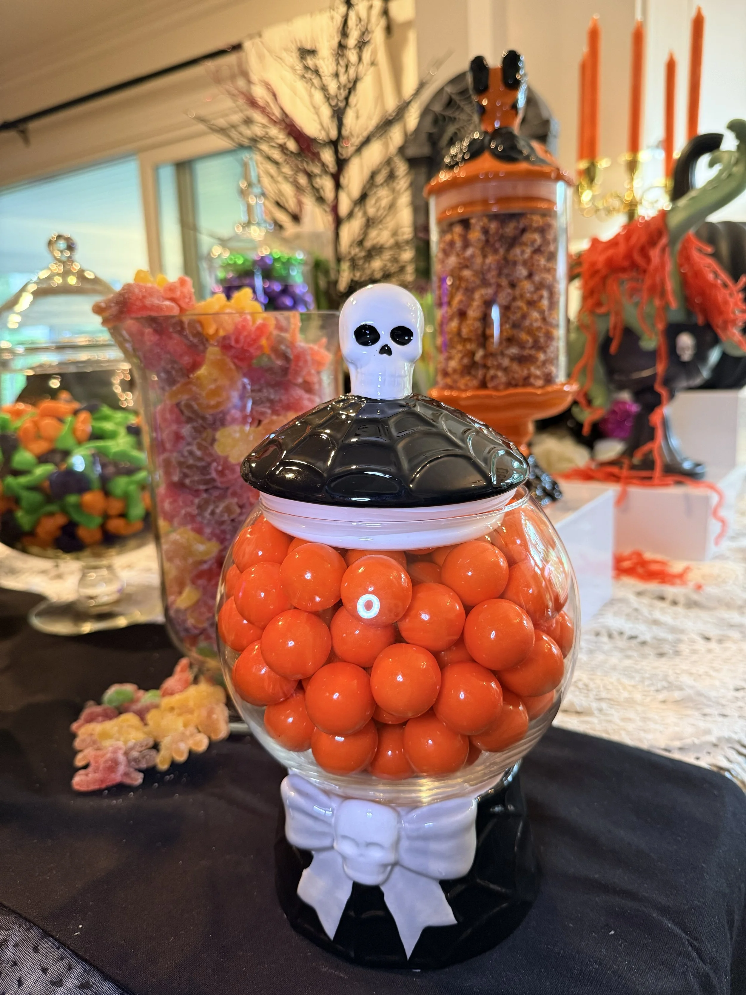 A glass jar filled with orange spherical candies, topped with a black and white skull-shaped lid, placed on a table among decorated Halloween treats and decorations.
