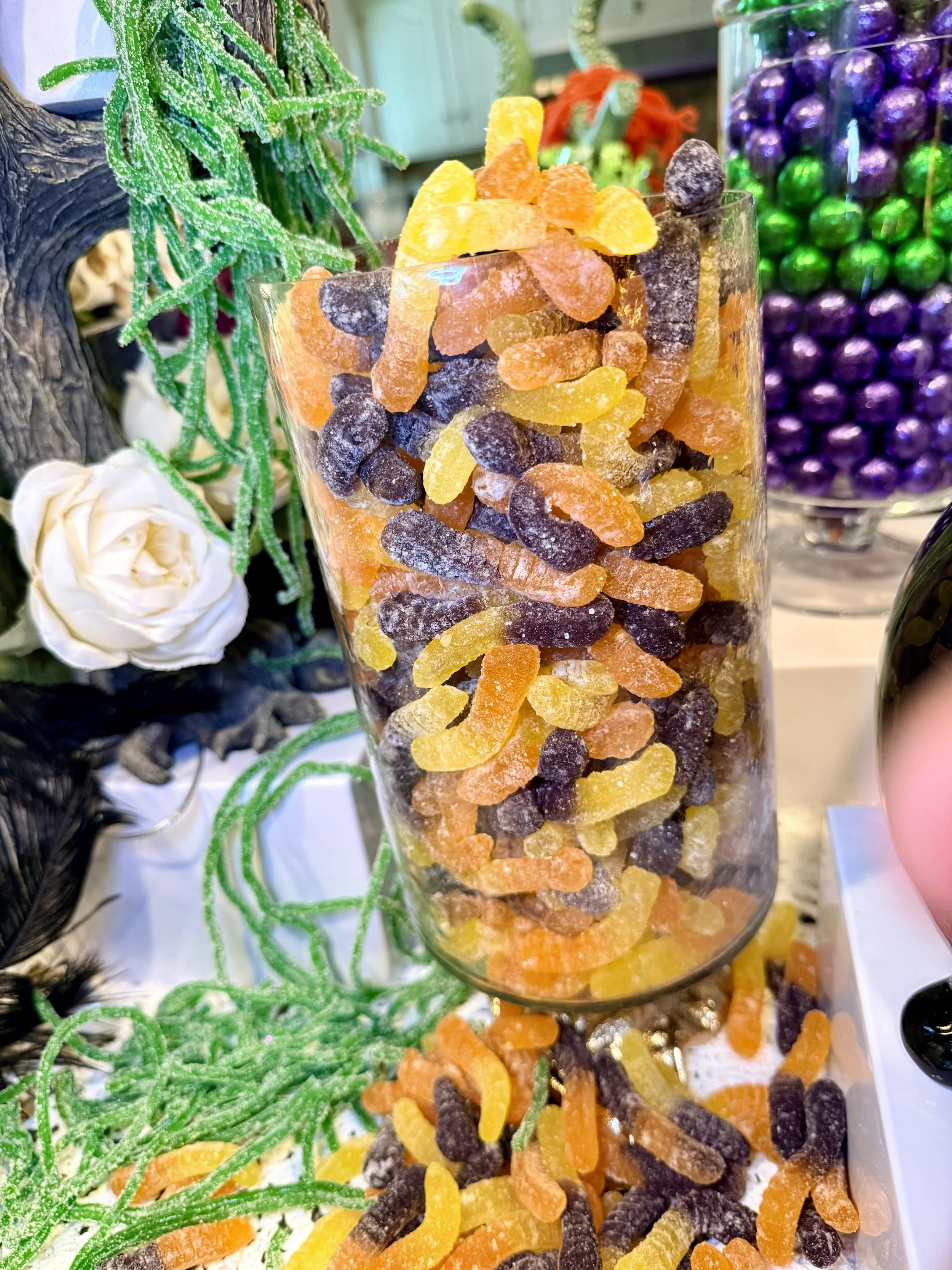 A tall glass filled with colorful sugar-coated gummy candies in orange, yellow, purple, and black, with some candies spilled around the base. In the background, there are decorative objects including a white rose, green textured materials, and a cont