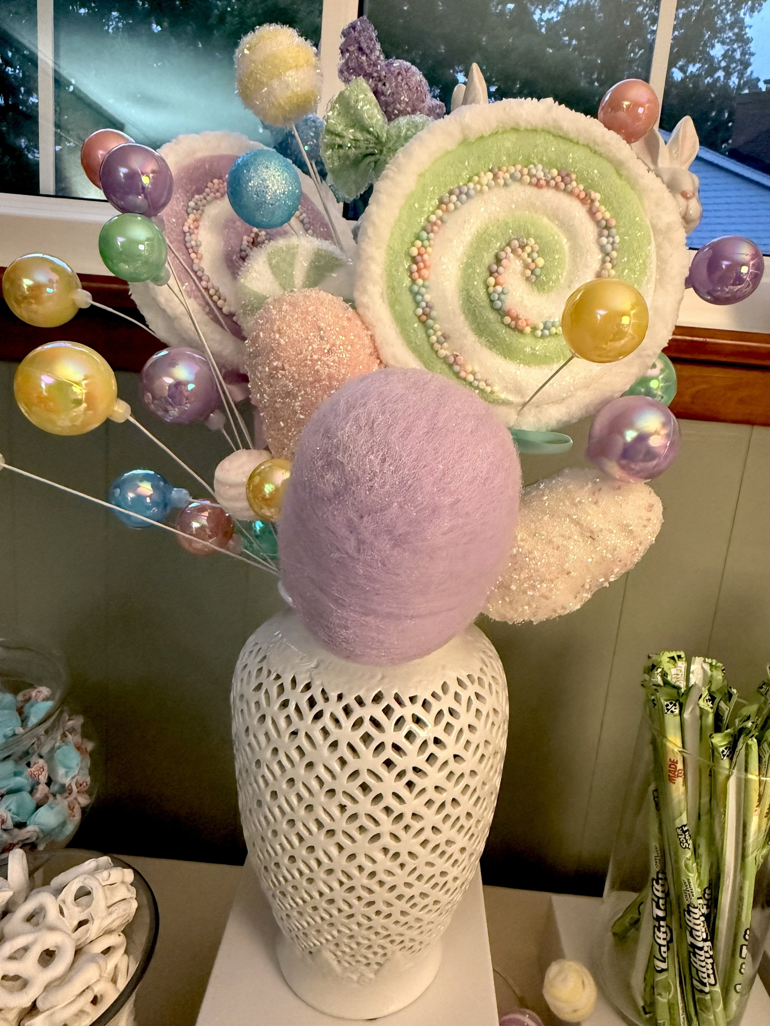 Decorative floral arrangement featuring large pastel-colored swirled lollipops with candy-covered spheres and cotton-like textured balls in pale pink, yellow, lavender, and green, displayed in a white decorative vase.