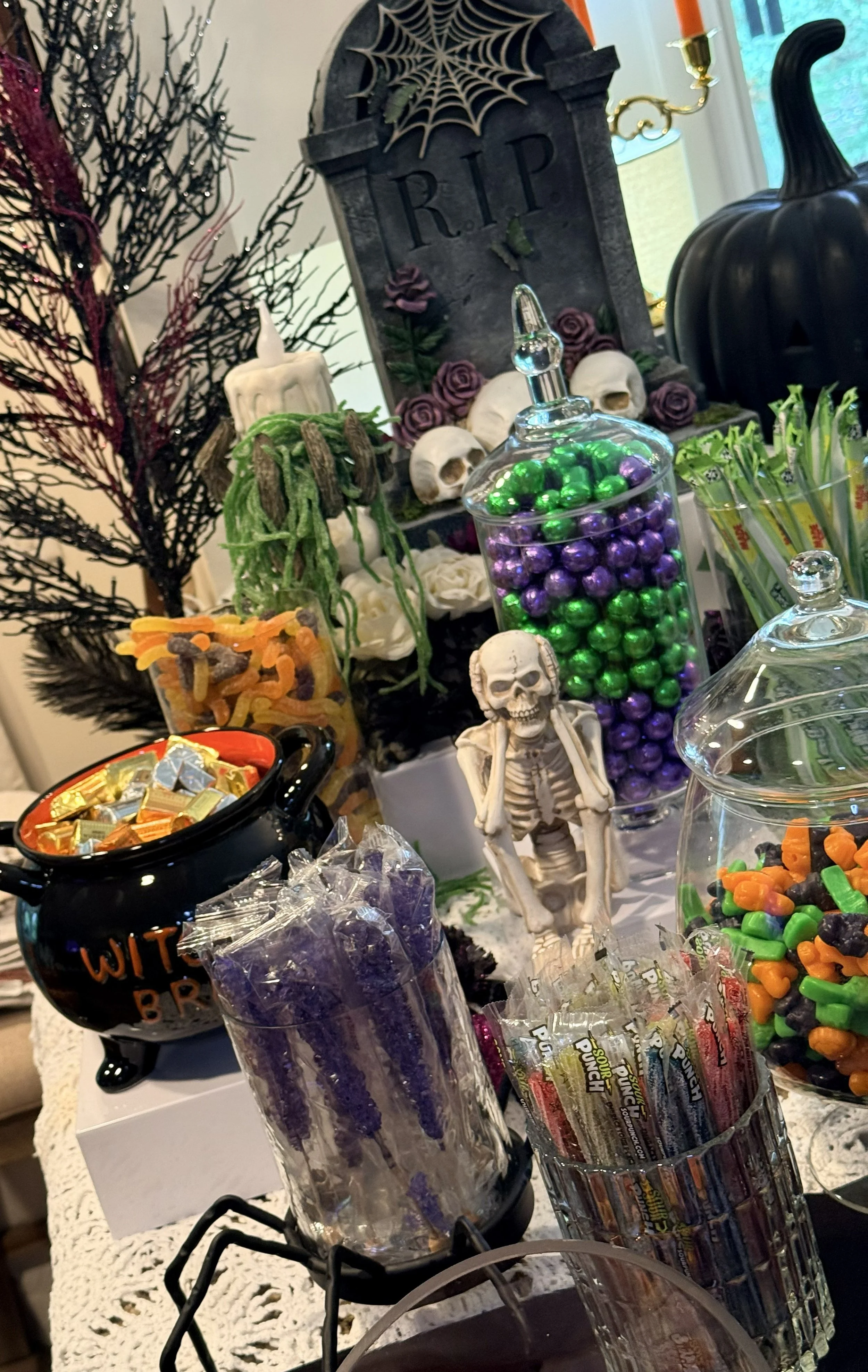 Halloween-themed candy display with black pumpkin, skeleton figurine, tombstone decoration, spider web, skulls, and various candies, including chocolates and gummy worms, set on a decorated table.