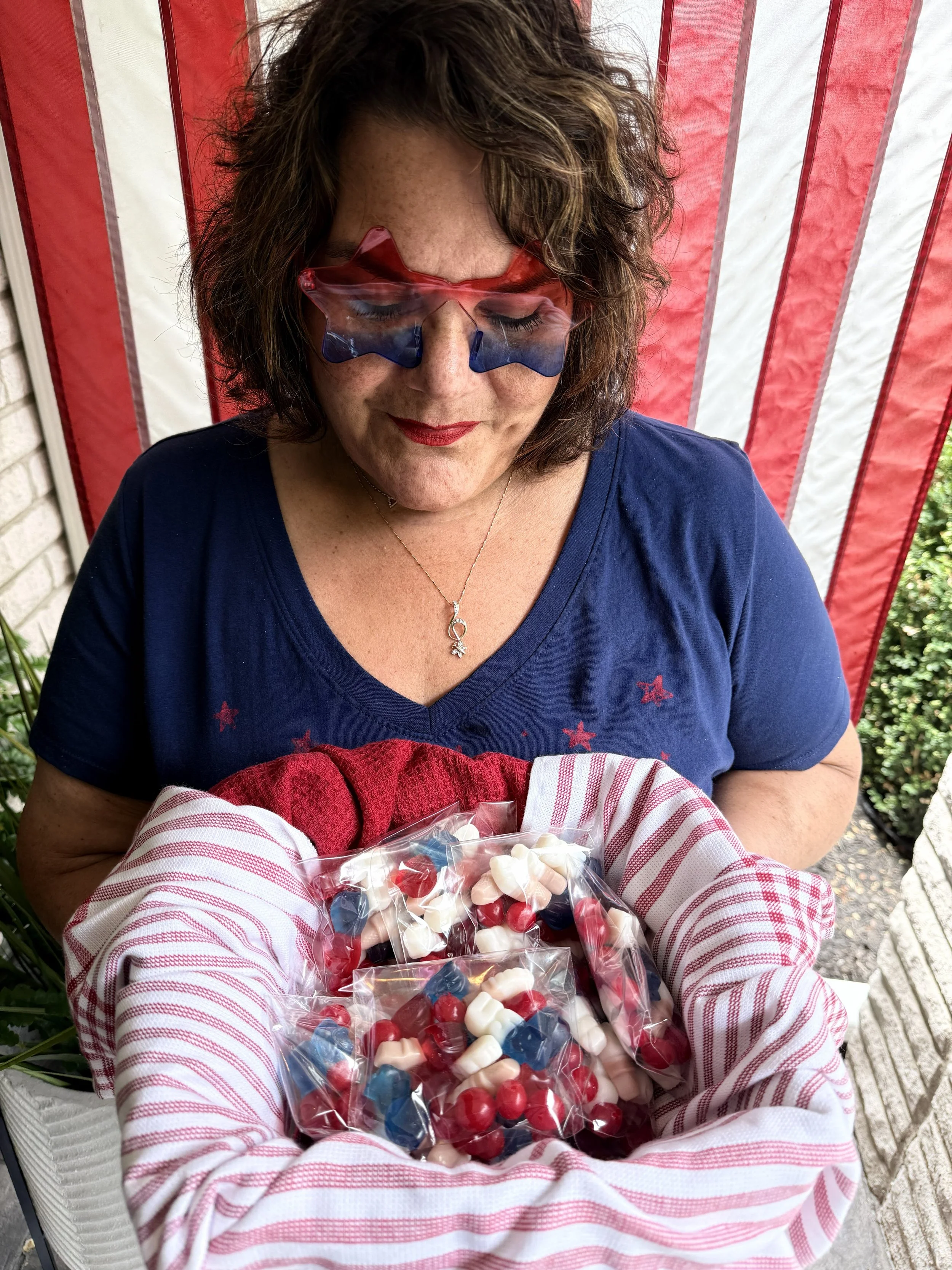 A woman wearing patriotic star-shaped glasses with red, white, and blue colors, smiling with her eyes closed, sitting behind a plastic bag filled with red, white, and blue candies, with an American flag-themed background.
