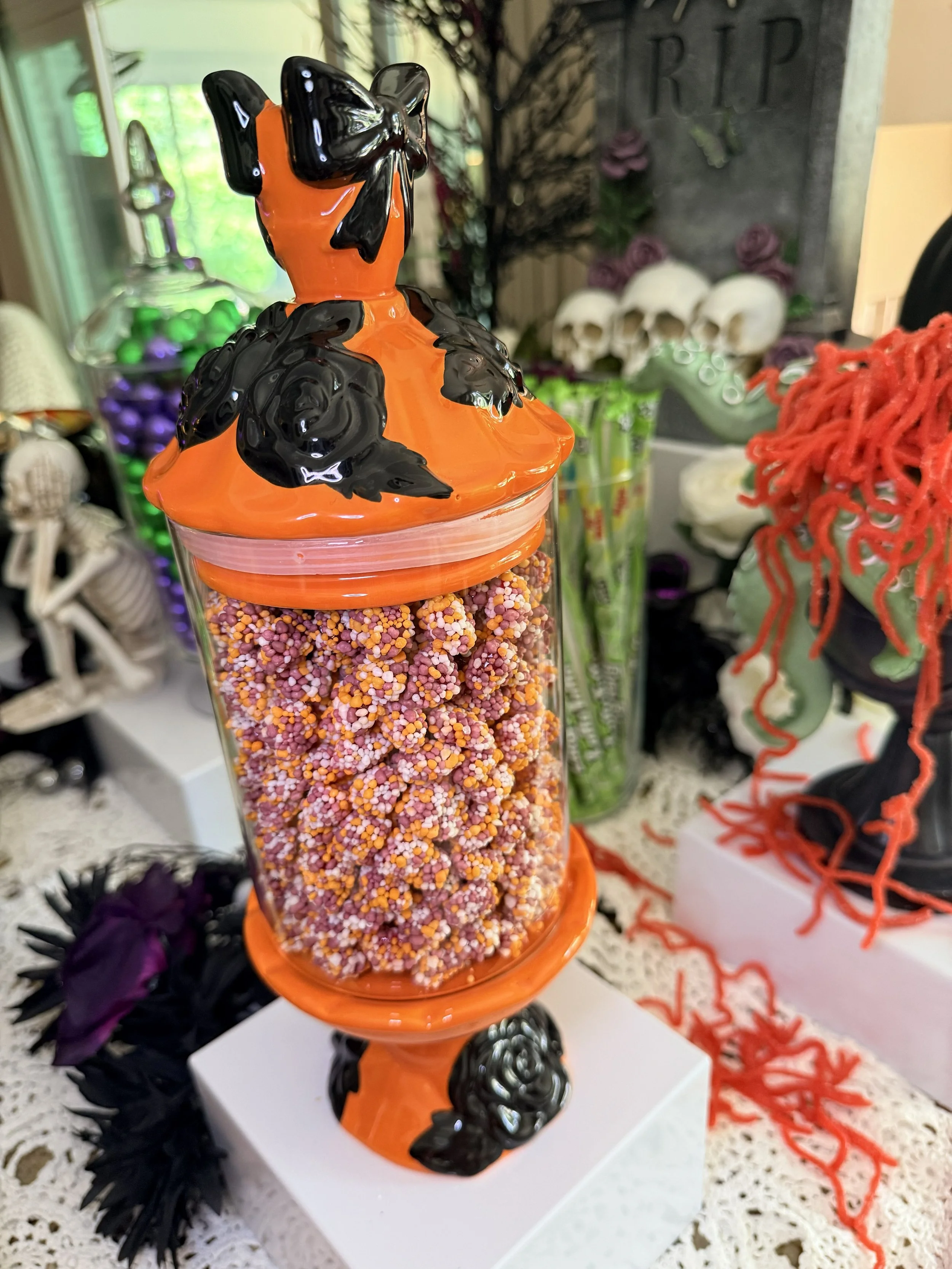 Halloween themed candy jar with orange lid, black bow decoration, and black rosettes, filled with colorful mini sprinkles, on white display table with other costumes and decorations in the background.