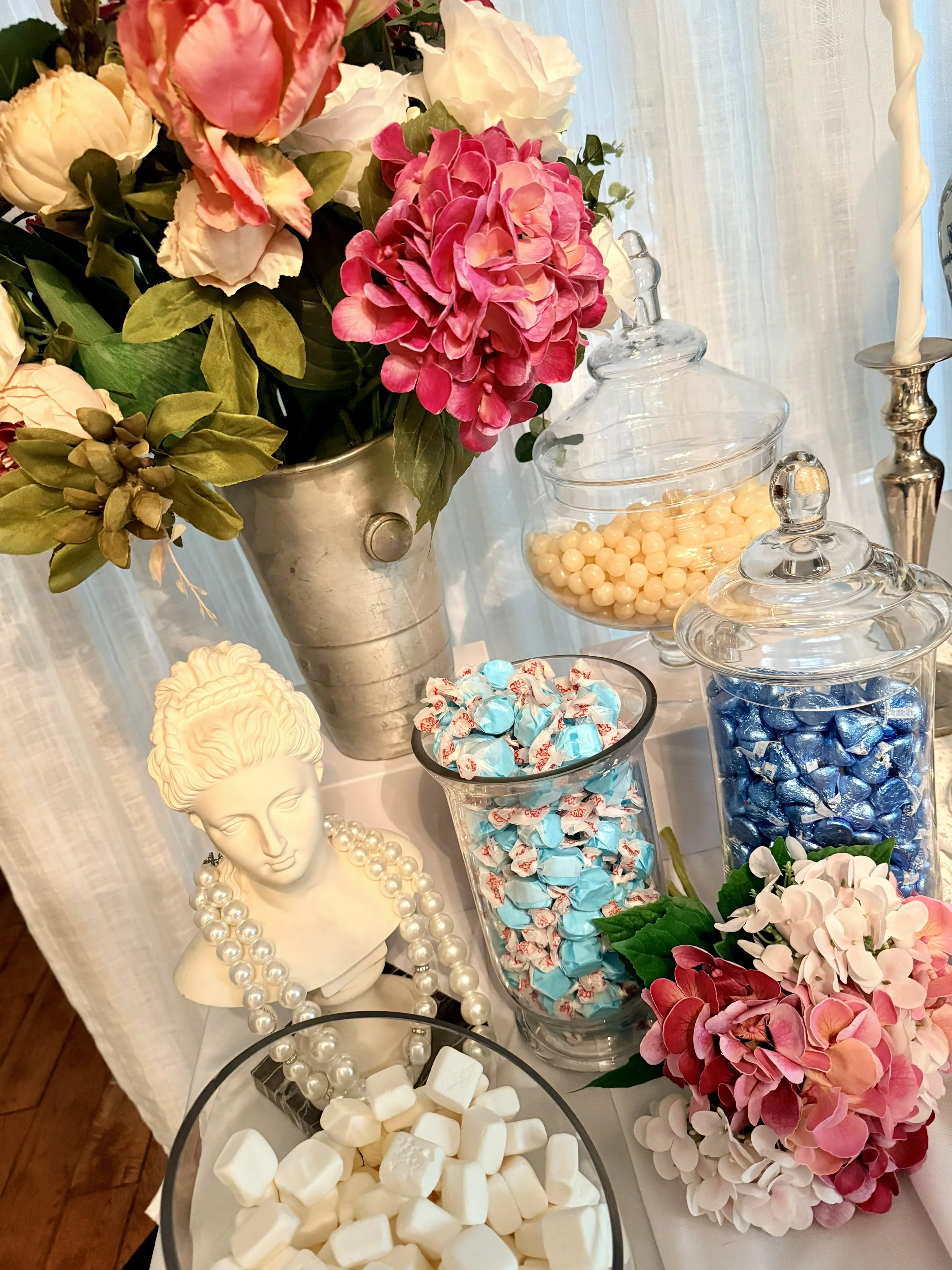 A display of pink and white hydrangea flowers in a silver vase, with glass jars containing candies and marshmallows, a decorative head bust with a pearl necklace, and a candlestick with a twisted white candle in the background.
