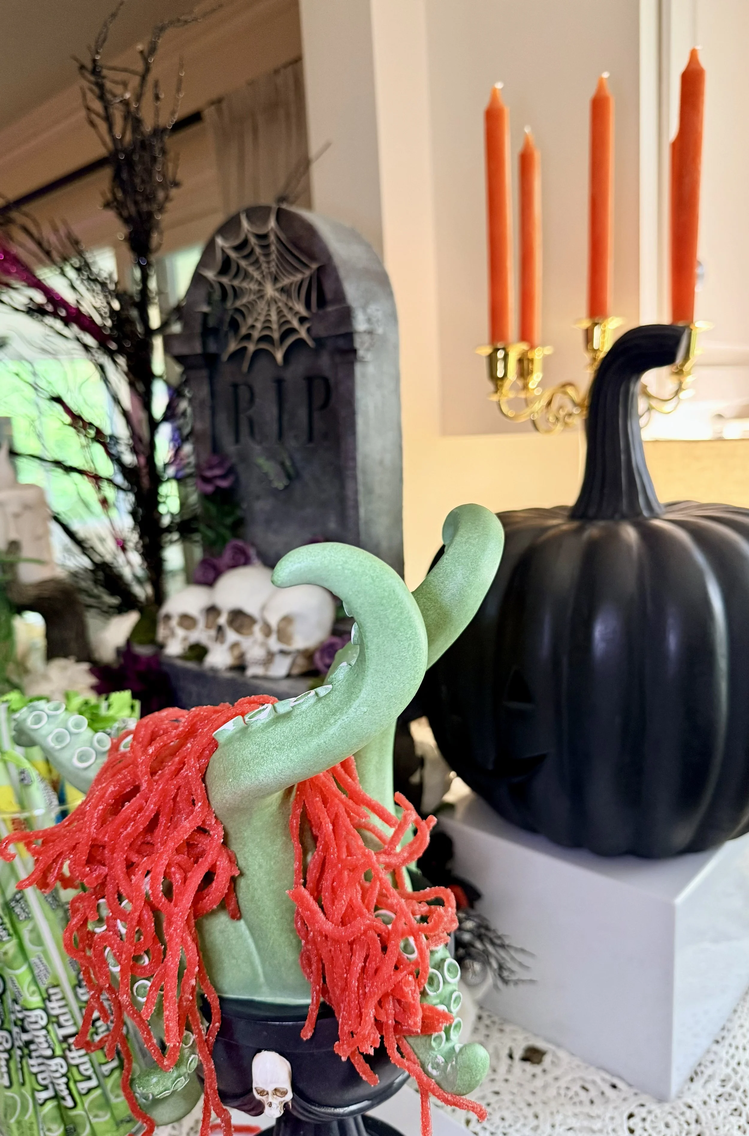 Decorative Halloween scene with a black pumpkin, red and white candles, a skull, a spider web on a gravestone, and a green dragon figure with red yarn details.