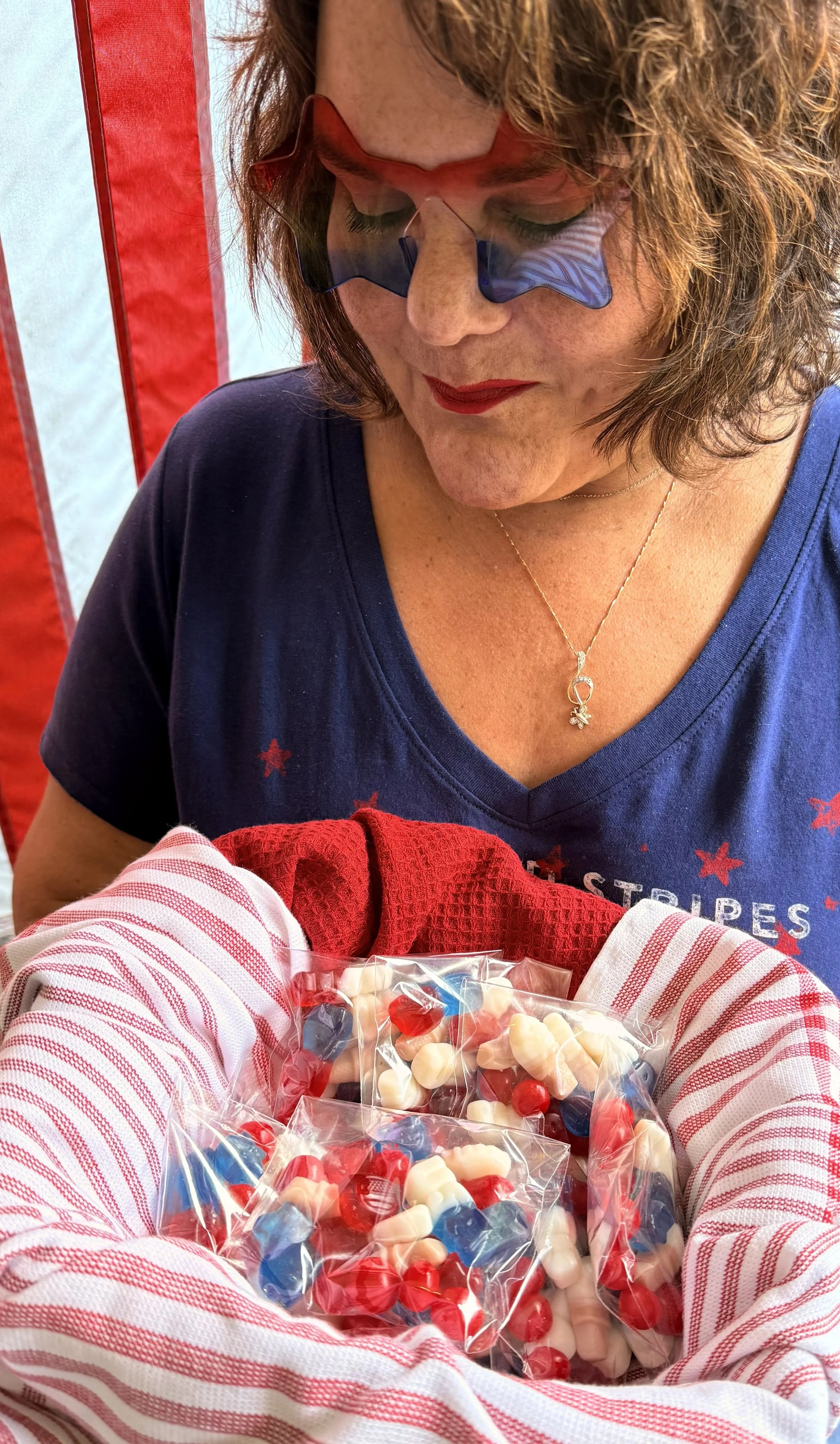 A woman with curly hair wearing sunglasses and a blue T-shirt is looking down at a bag of white, red, and blue gummy candies held by a child in a red and white striped shirt.
