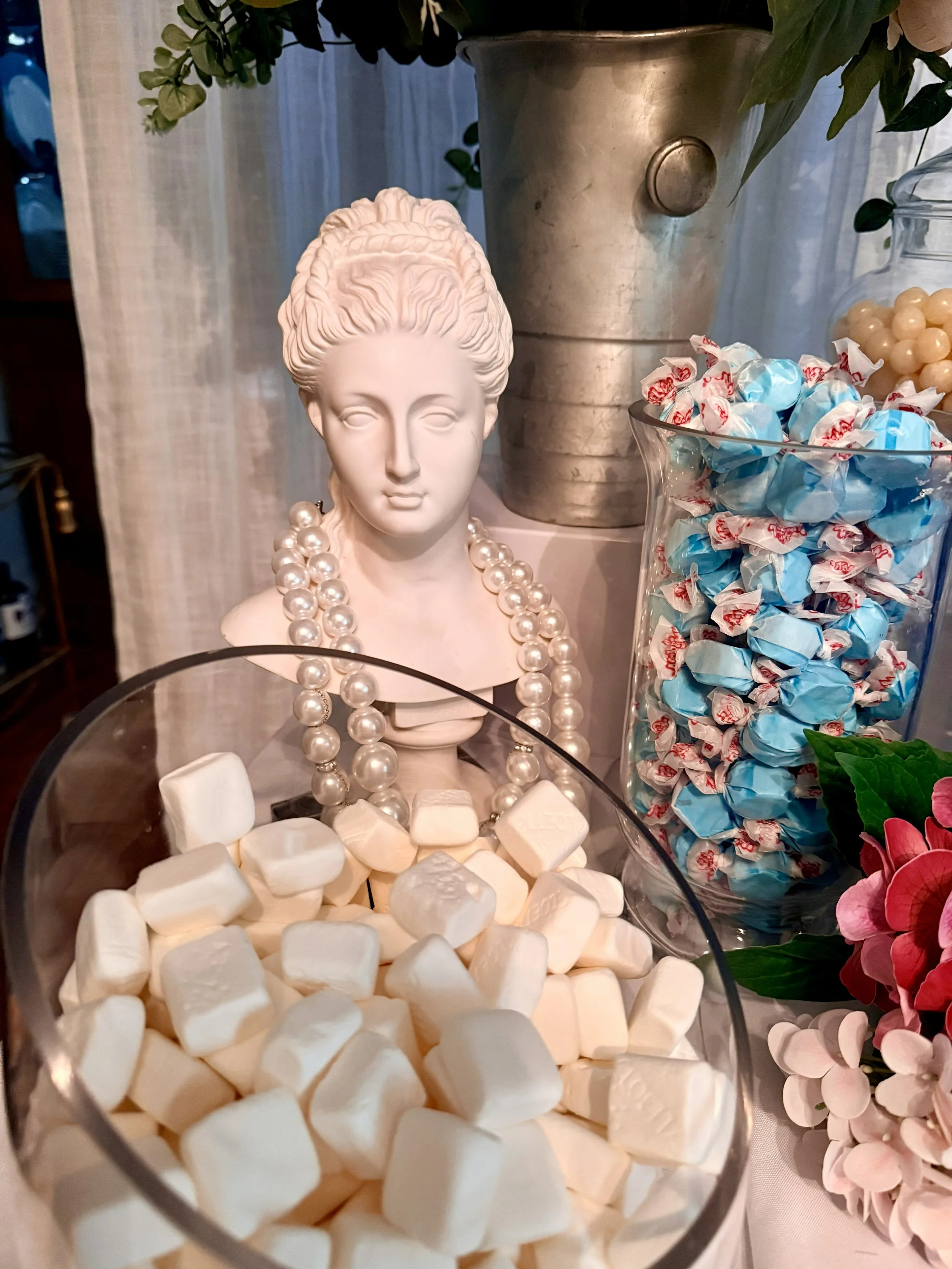 Decorative display featuring a white bust sculpture wearing pearl necklaces, surrounded by candies, flowers, and a metal vase.