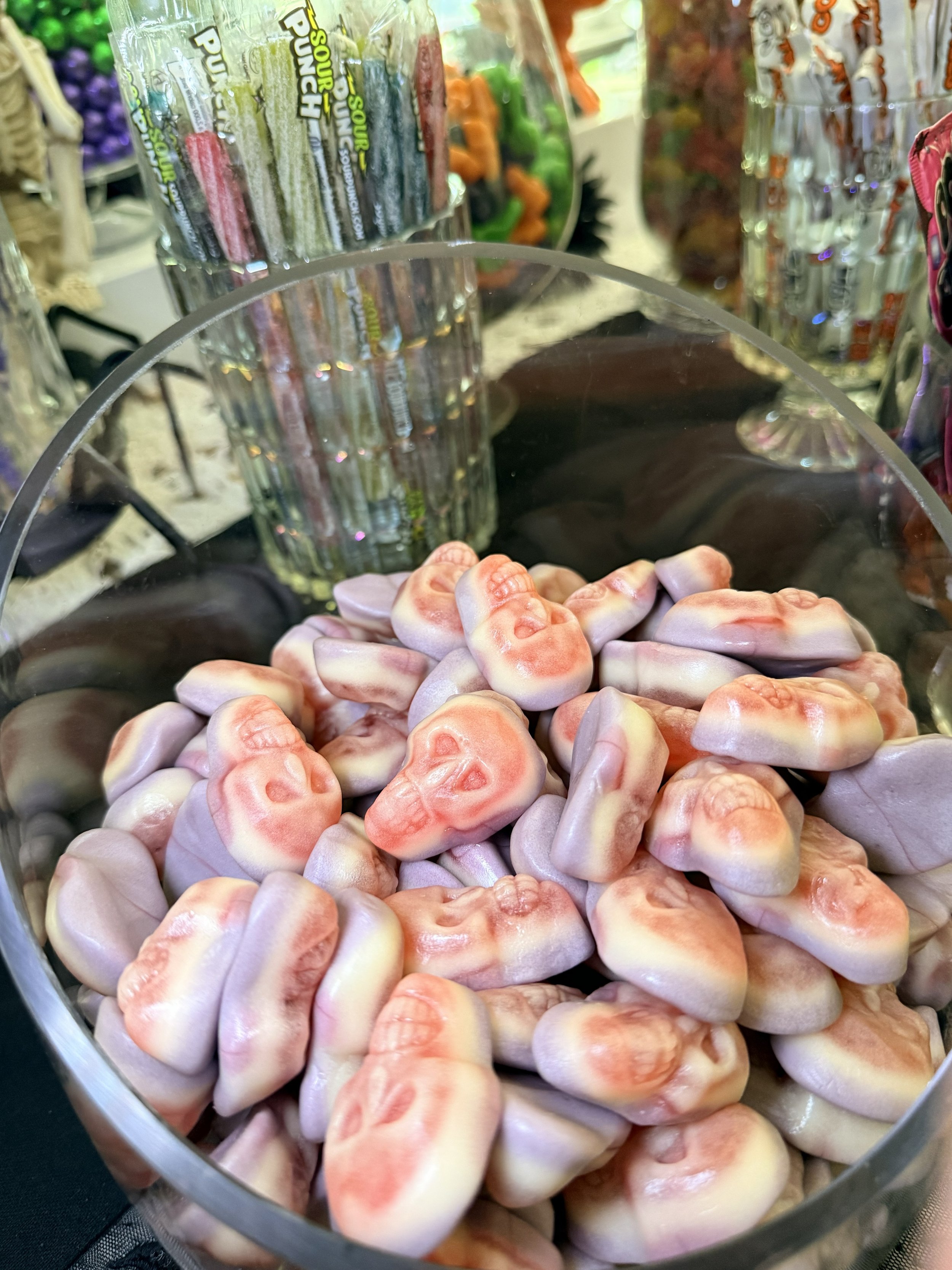 A clear container filled with pink and white carved rocks, with some green, purple, and orange objects and glassware with colorful plastic items behind it.