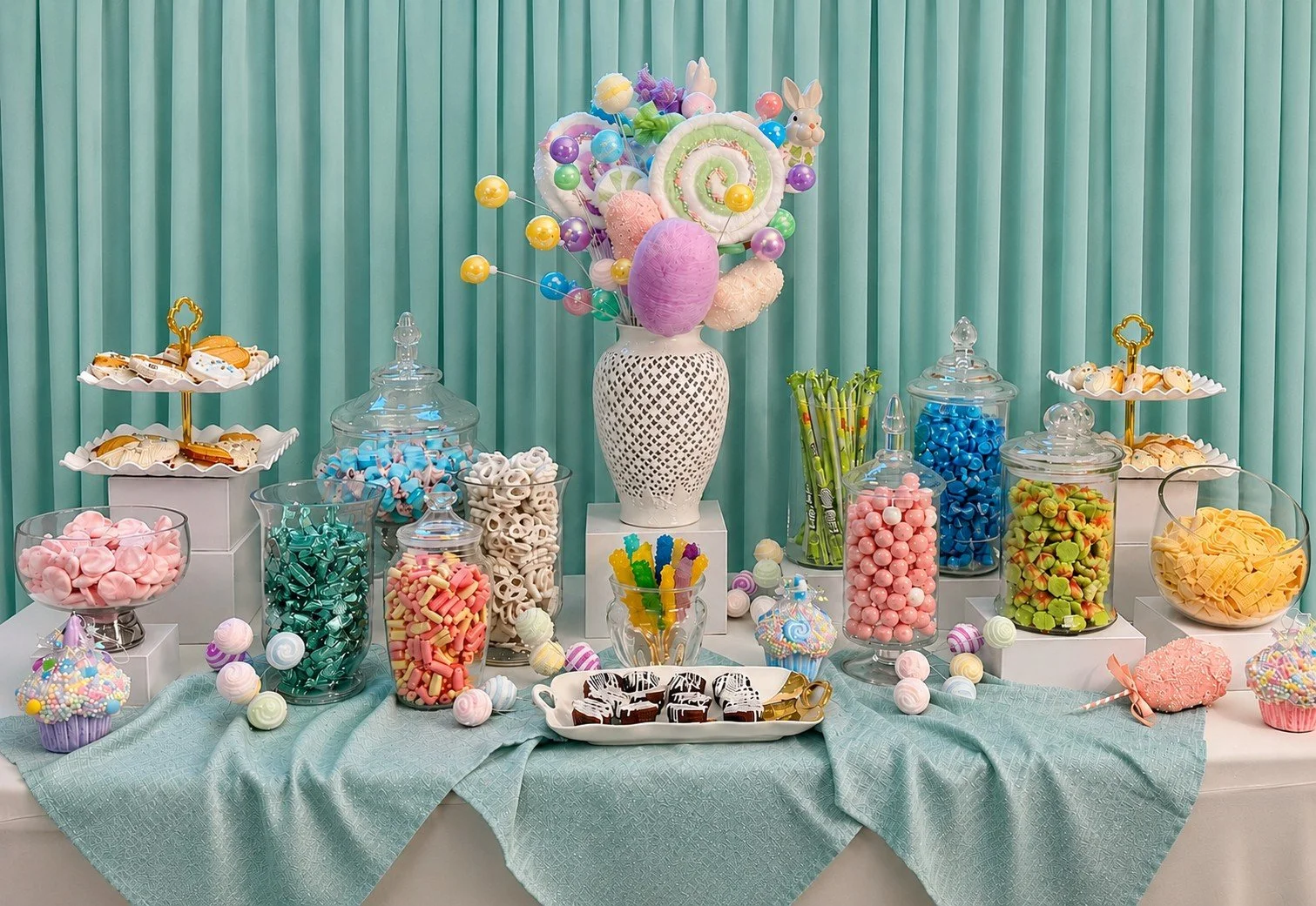 Colorful candy and dessert table with a large white vase of pastel-colored cotton candy, surrounded by jars of sweets like gumdrops, gummies, and chocolate, topped with cupcakes and decorated for a celebration.