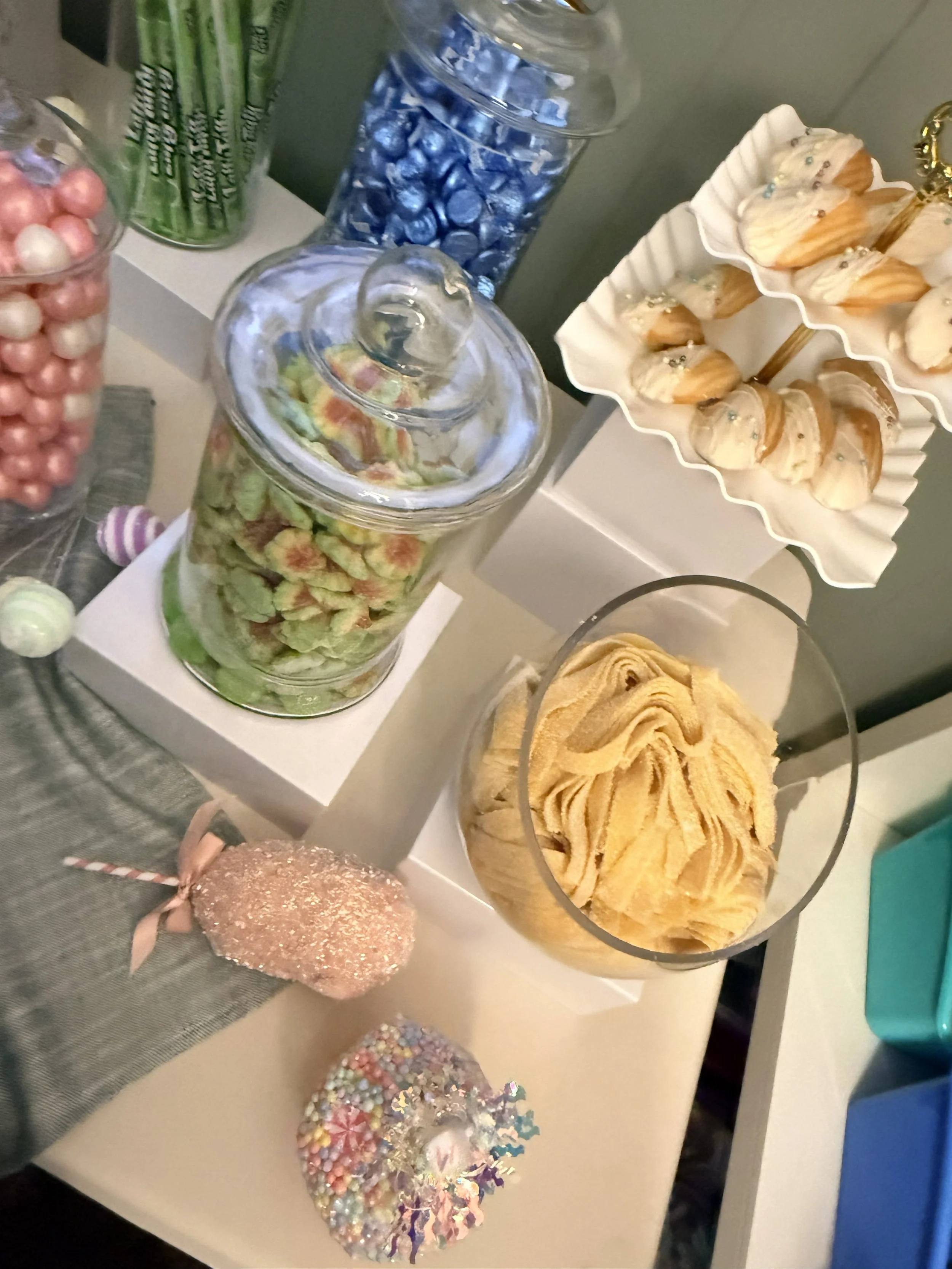 Assorted candies and desserts on a table, including colorful pastel meringues, cupcakes with white frosting and silver sprinkles, blue and pink candies in jars, and confetti decorations.