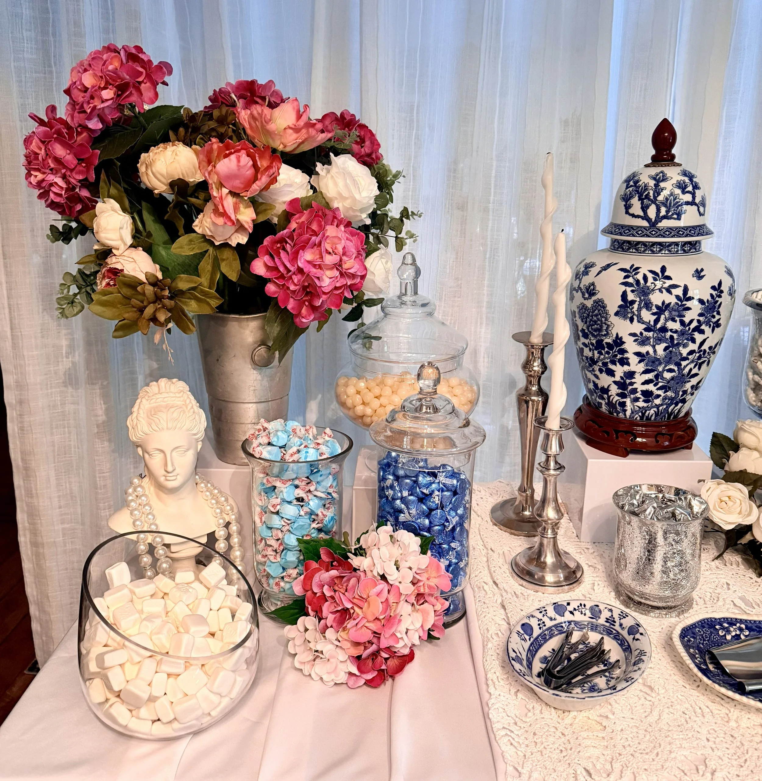 A floral arrangement of pink and white peonies and hydrangeas in a silver vase on a table with decorative glassware and porcelain dishes with blue floral designs.