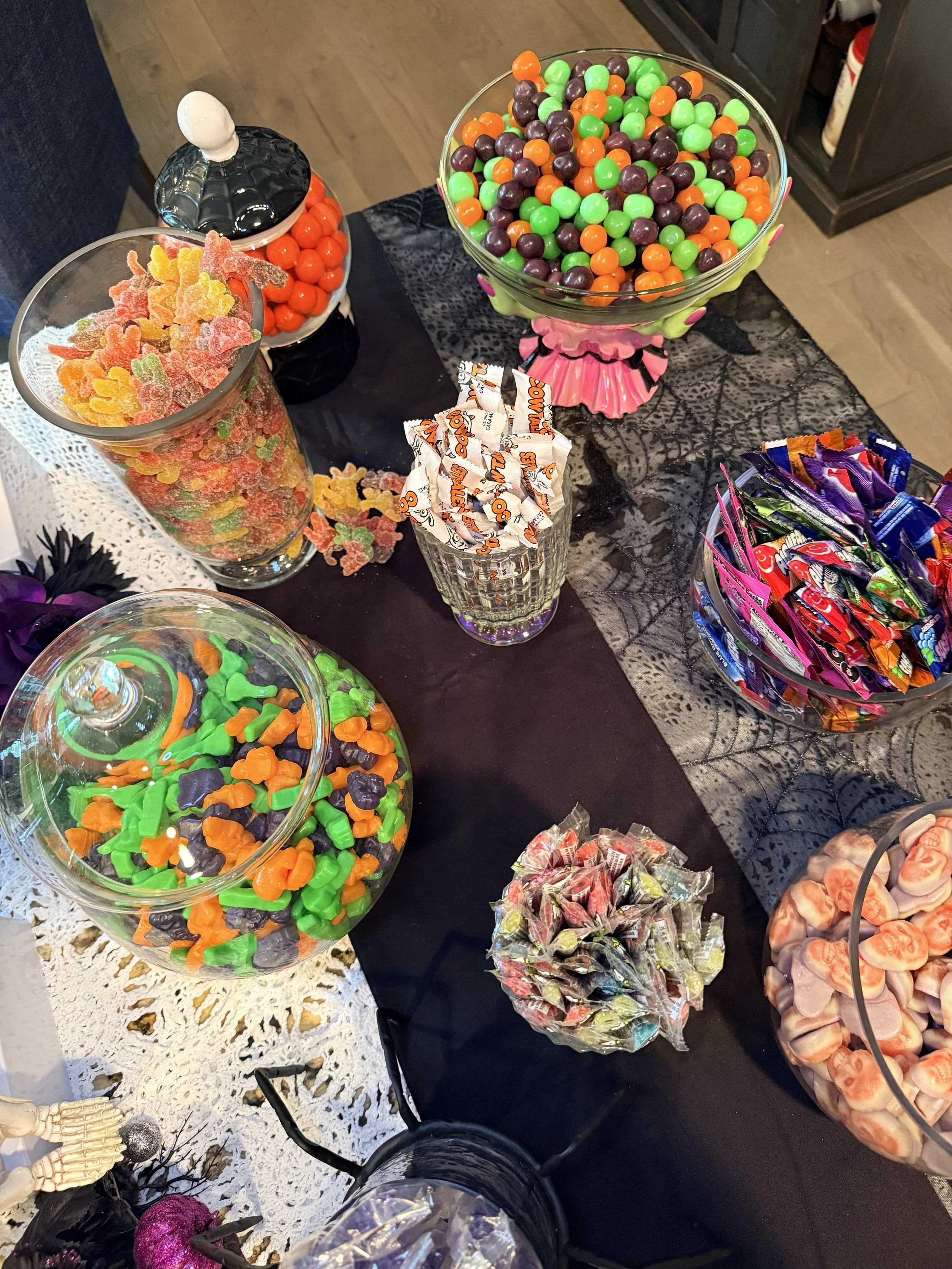 Assorted candies in bowls, including gummy worms, gummy bears, M&Ms, Reese's, wrapped candies, and lollipops, arranged on a black tablecloth.