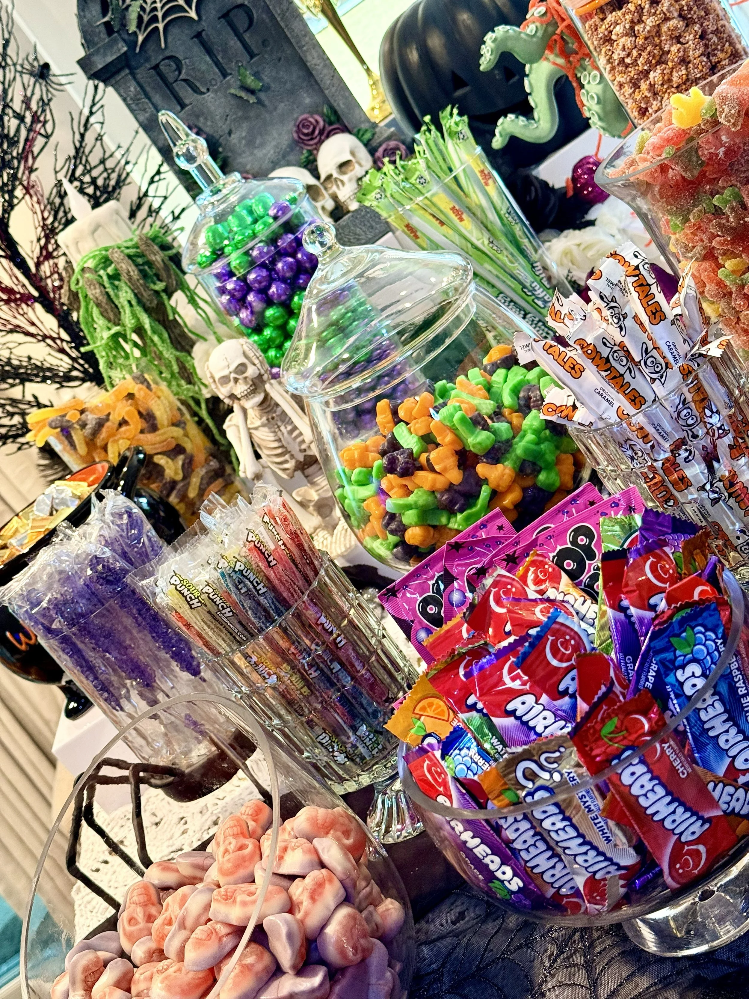 Glass jars filled with Halloween-themed candies, including green and purple chocolates, gummy worms, and other colorful candies, with candy packages and Halloween decorations such as skulls, skeletons, and a spider web in the background.