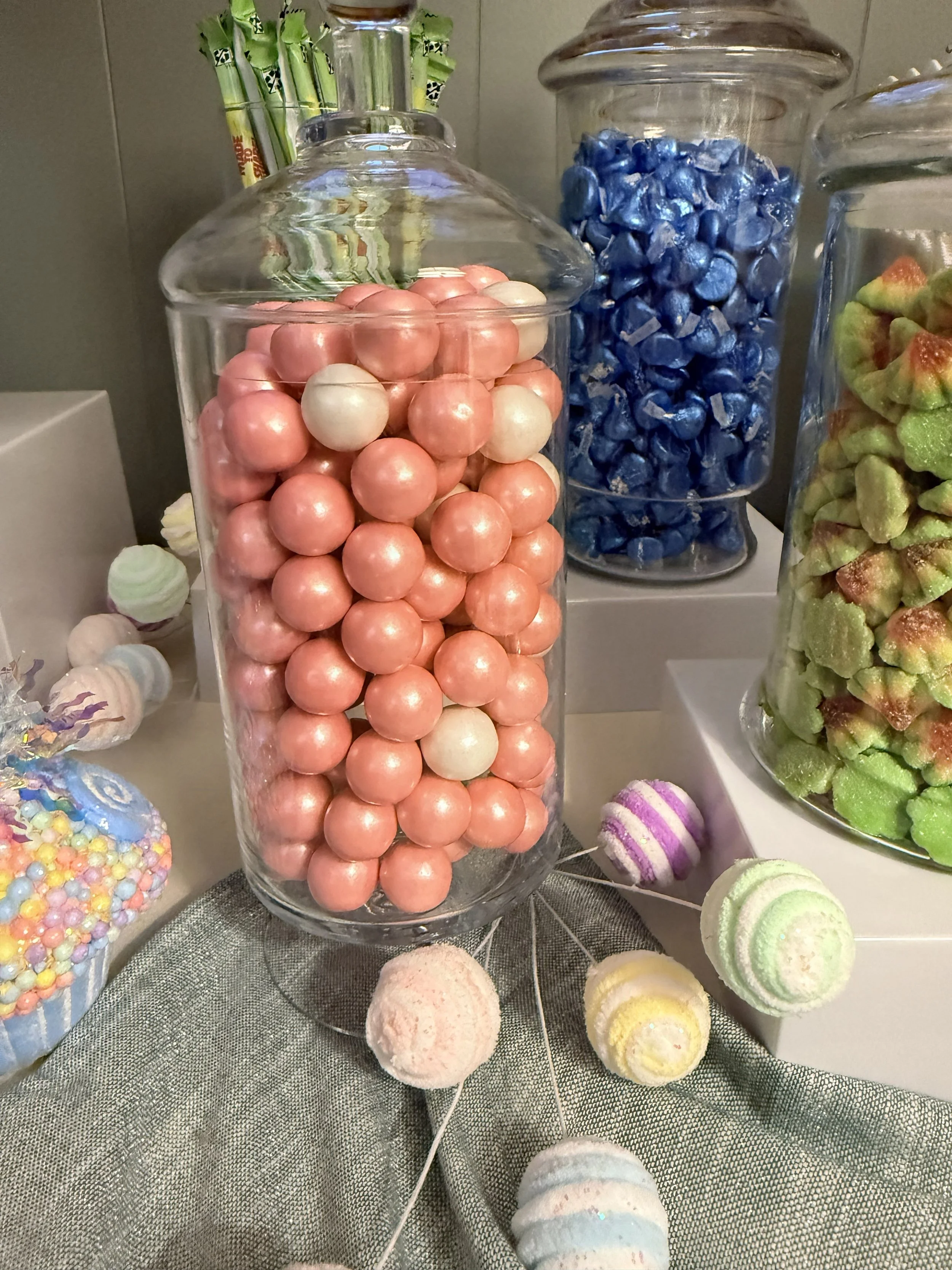 Assorted colorful candies including pink and white spherical candies in a glass jar, blue wrapped candies in another jar, and various pastel-colored candies and confections on a gray fabric surface.