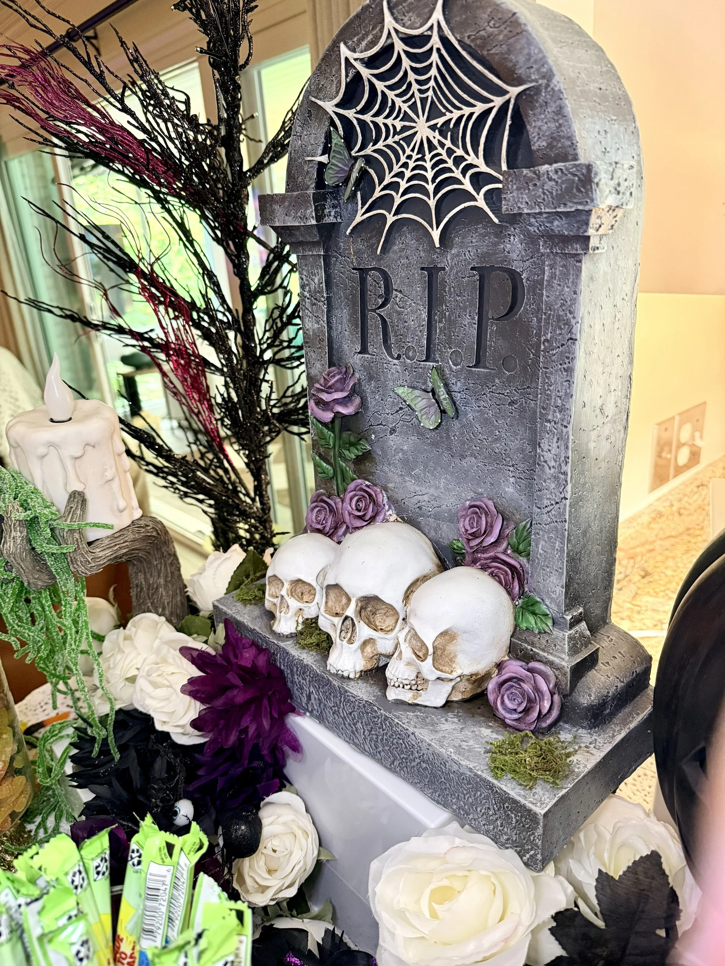 A Halloween-themed display with a gray stone-like tombstone that has a spider web and the initials 'R.I.P.' carved into it. The display is decorated with four artificial skulls, purple roses, white and black flowers, and various decorative elements i