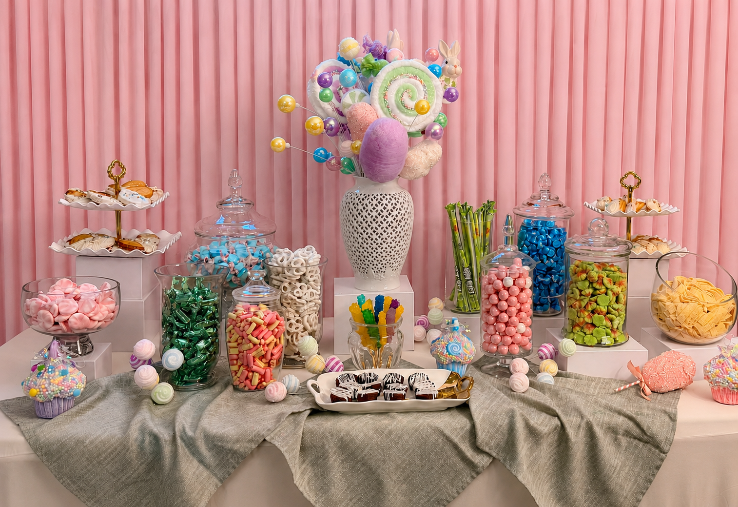 Sweet treat display with pink backdrop, featuring an arrangement of colorful candies, chocolates, cupcakes, and decorated desserts on a table decorated with a grey tablecloth.