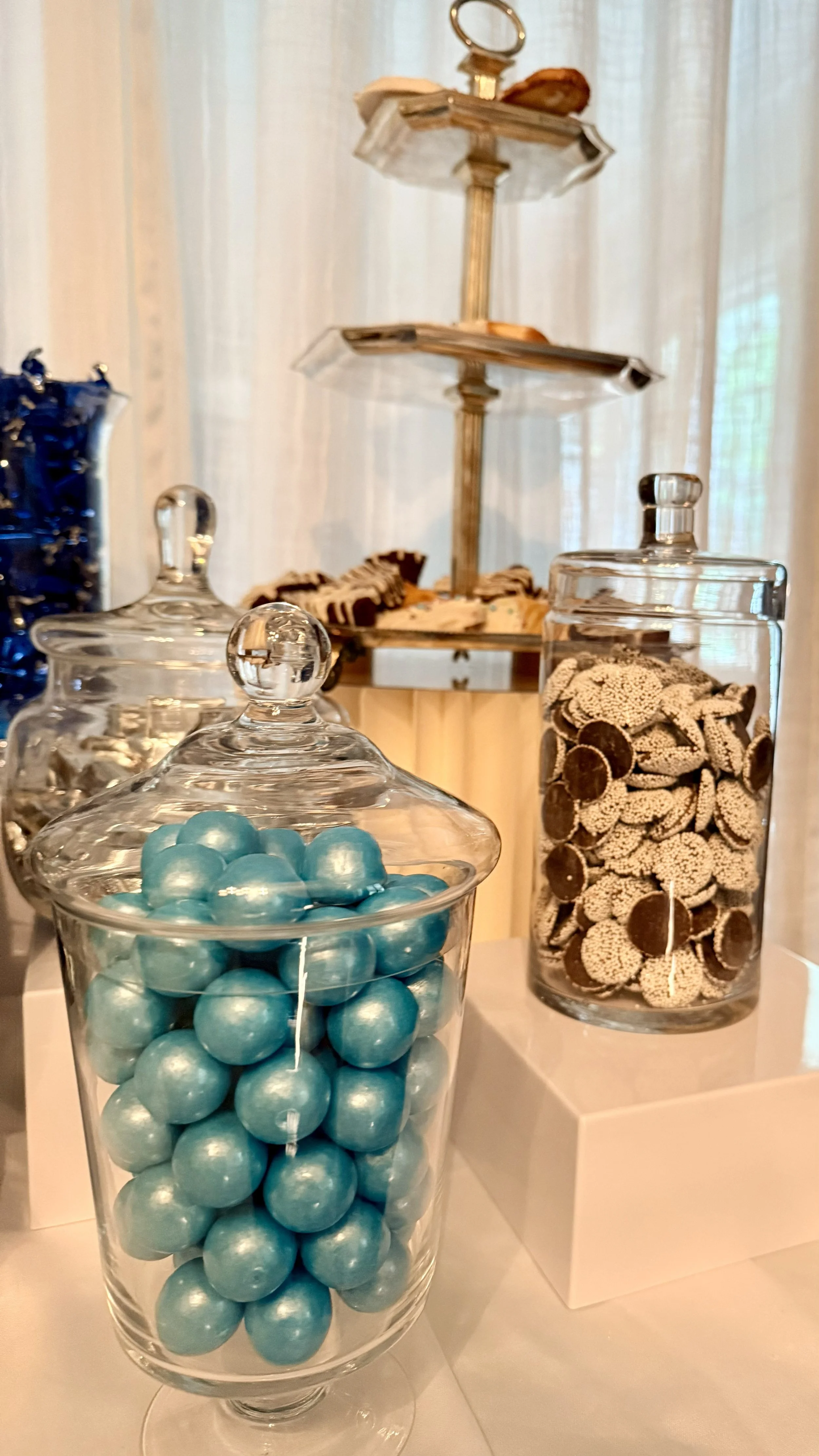 Glass jars filled with decorated candies, including blue spherical candies and beige candies with brown centers, along with a two-tiered tray holding additional treats in a decorative setting.