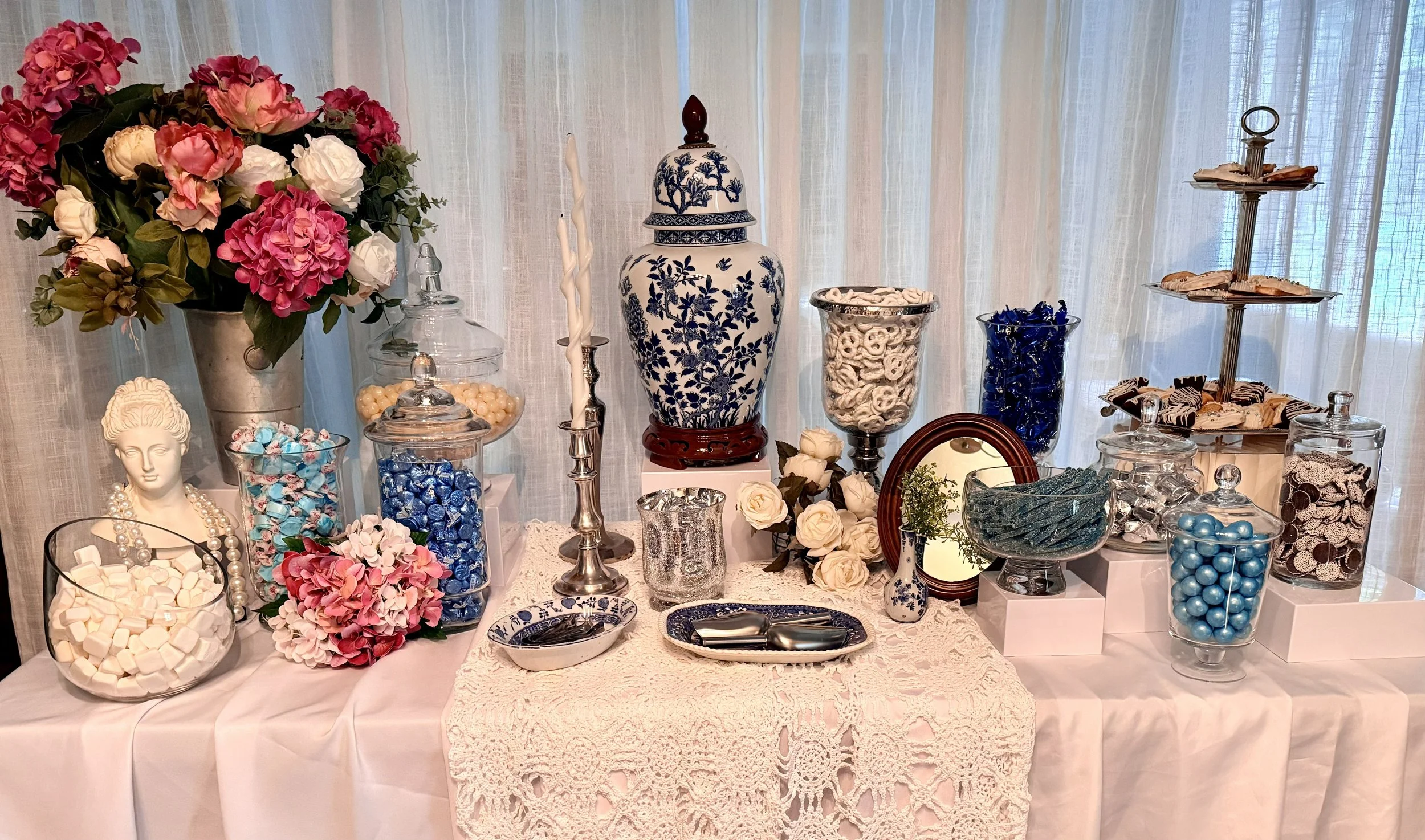 A table decorated with blue and white porcelain vases, jars, and plates, holding pastel-colored candies, chocolates, and flower arrangements, with a lace tablecloth and sheer curtains in the background.