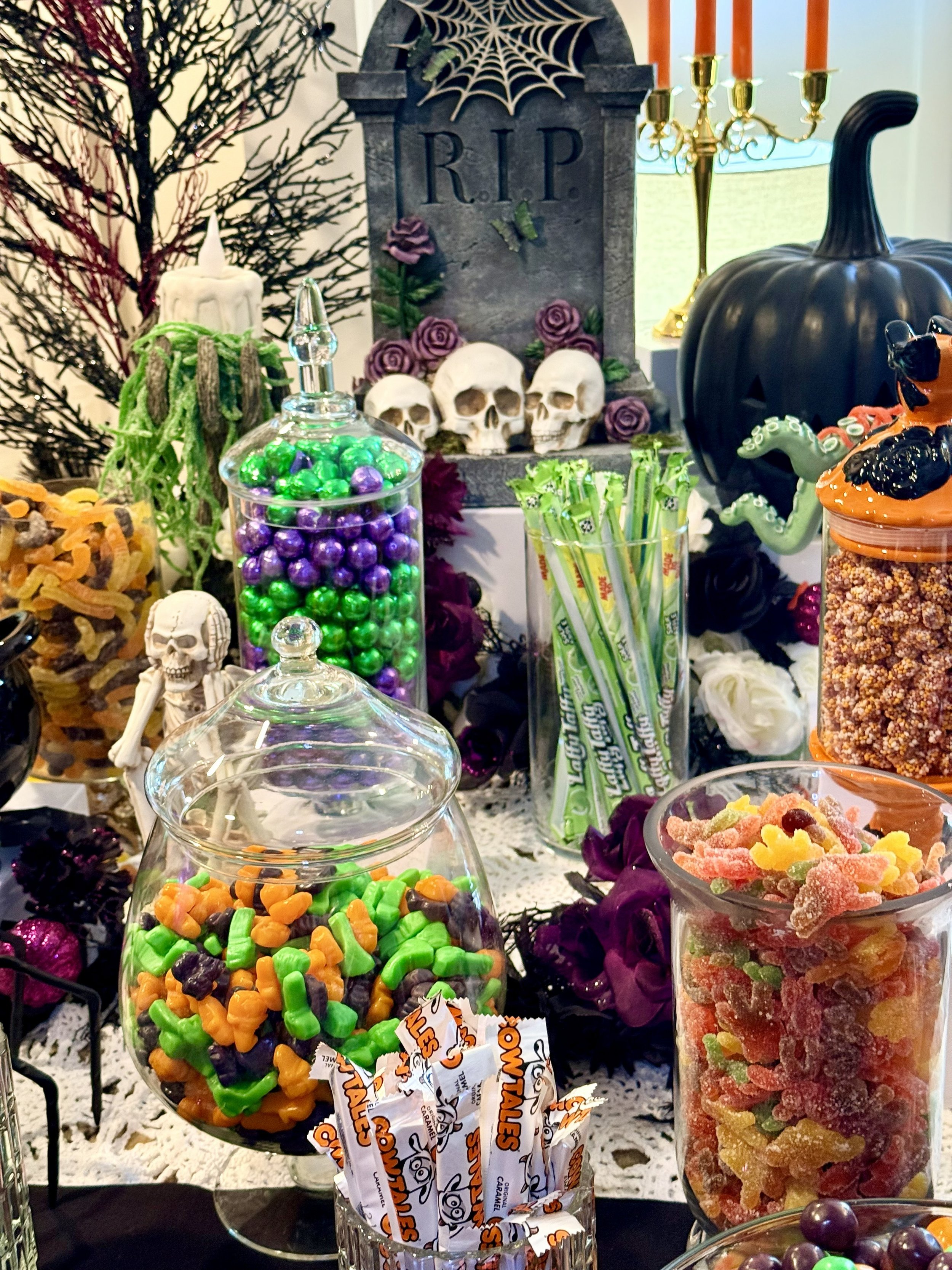 Halloween-themed candy display with colorful candies, skull decorations, black pumpkin, orange candlestick holder, and a gravestone with 'RIP' inscribed, accented by purple roses, a spider web, and a black tree in the background.