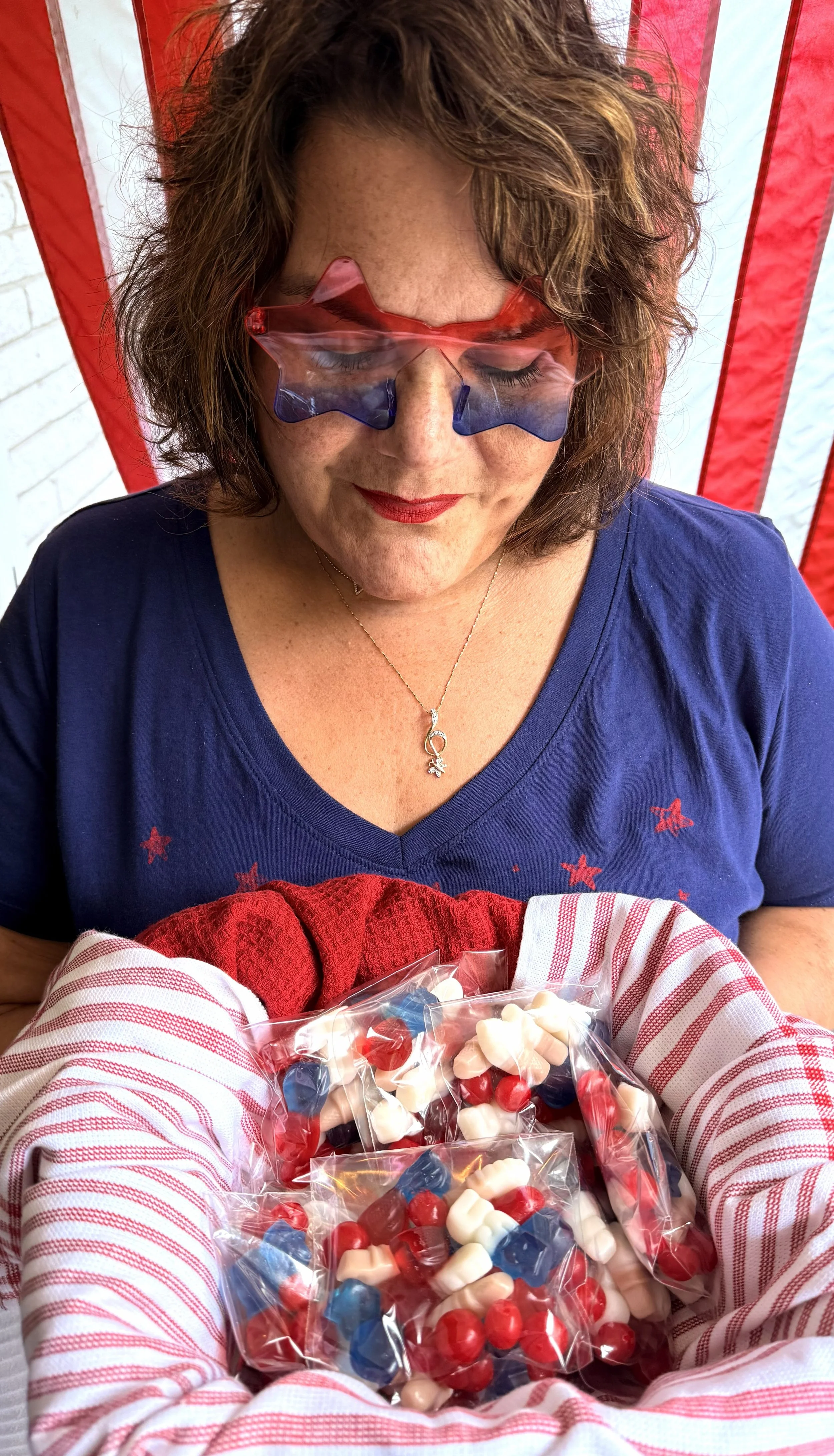 Woman with patriotic sunglasses opening a bag of red, white, and blue candies, dressed in patriotic-themed clothing, with a red and white striped cloth in front.
