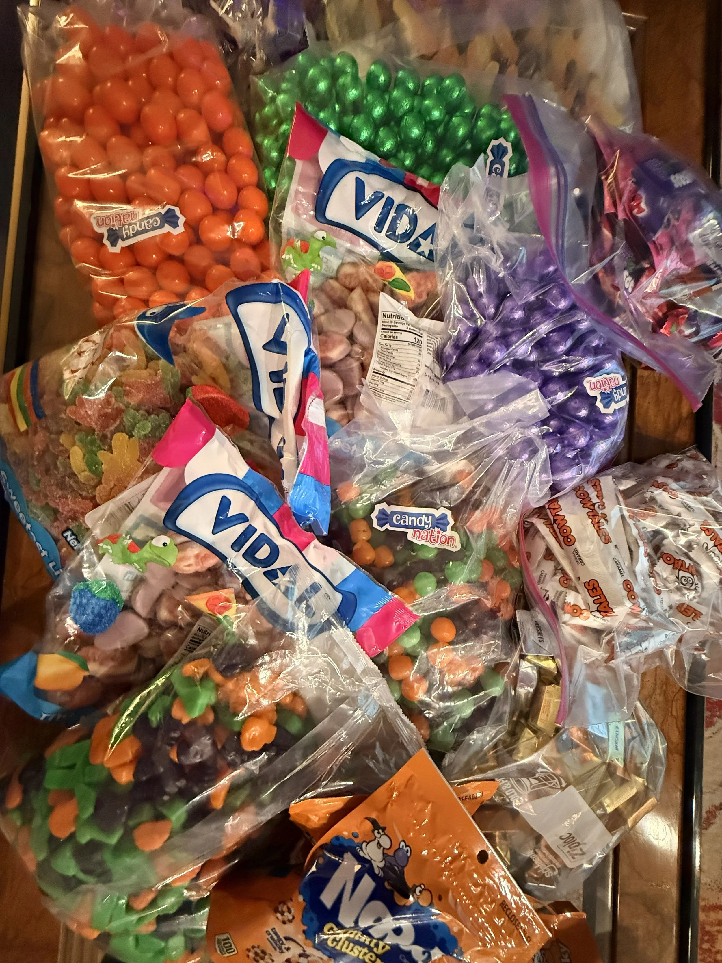 Assorted bags of colorful candies on a table, including green, orange, and purple candies, some with branding labels such as Vidia, Candy Nation, and others.
