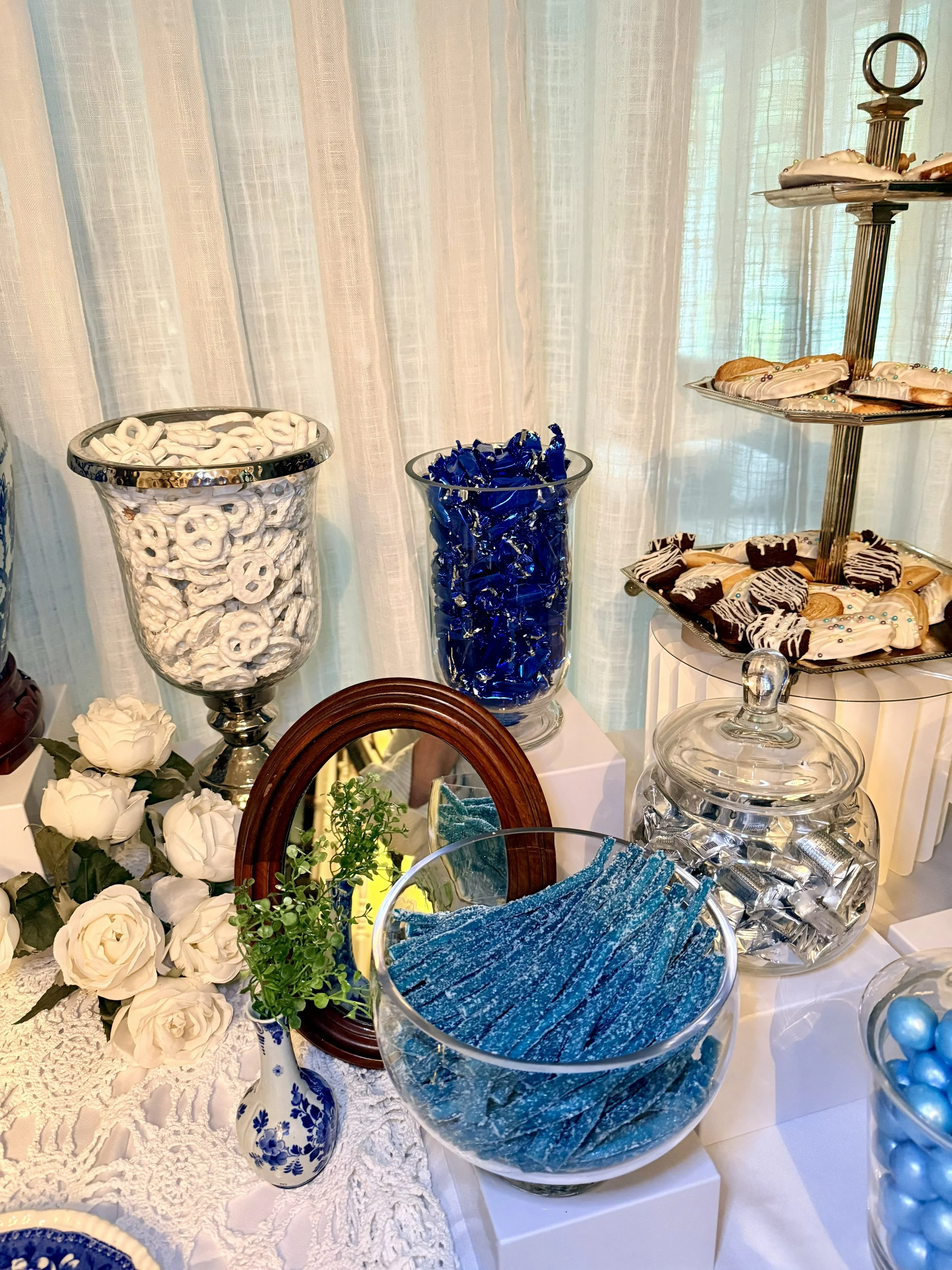 A display of various candies and desserts on a table, including pretzels, chocolate-covered treats, and blue candies in glass containers, with white roses and greenery as decoration.