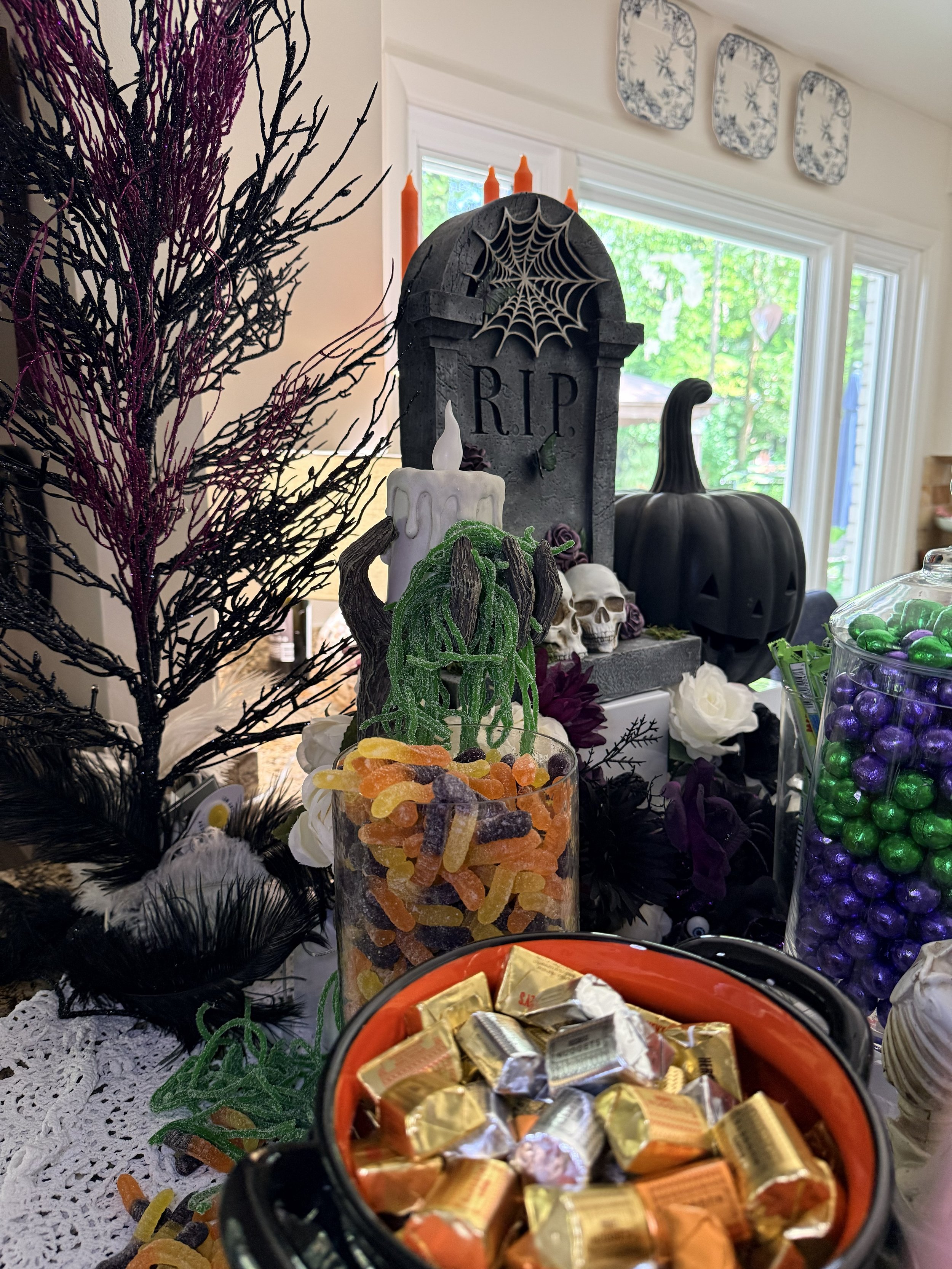 Halloween-themed table display with a black tombstone, orange candles, a black pumpkin, skulls, a candle with melted wax, decorative black and purple foliage, and bowls of candies including wrapped chocolates and gummy candies.
