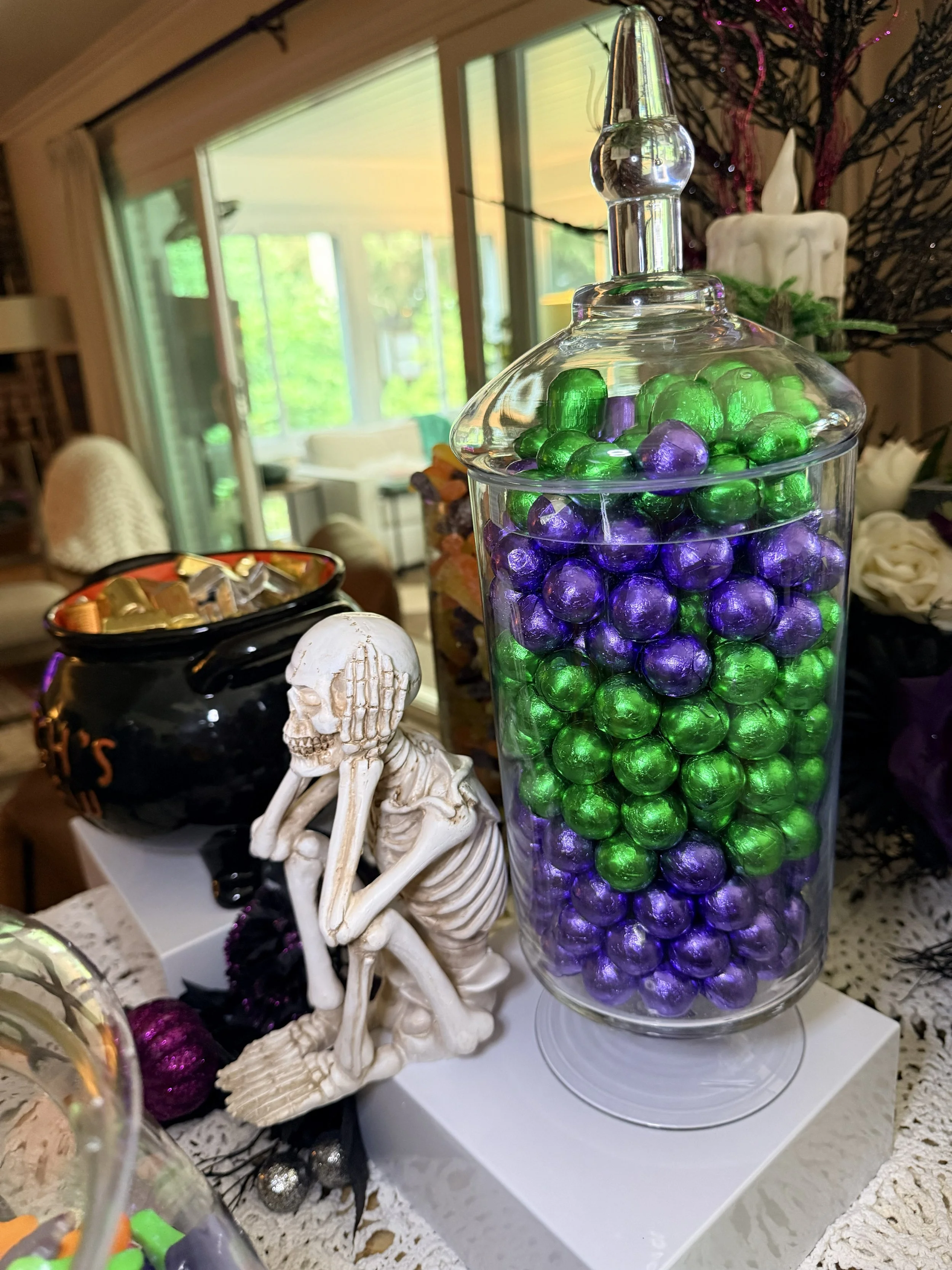 Glass jar filled with purple and green decorative glass balls on a table, with a skeleton figurine holding a skull and a Halloween-themed black cauldron with gold candy wrappers nearby, in a room decorated for Halloween.