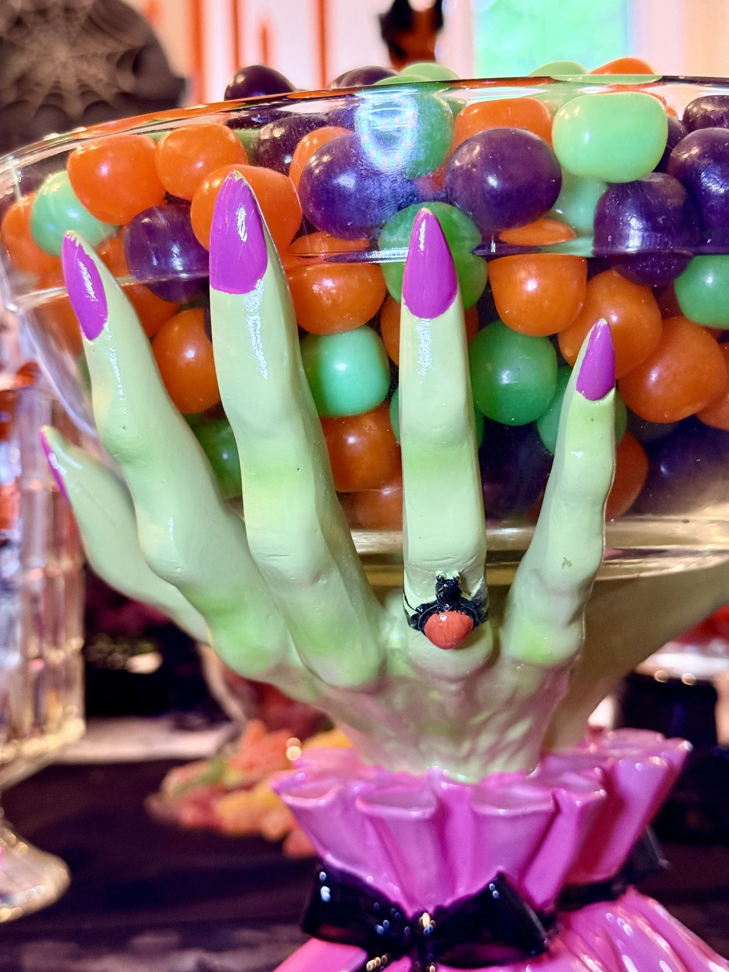 Decorative glass bowl filled with colorful plastic candy gumballs in green, orange, and purple, with a small ladybug figurine on the hand holding the bowl.