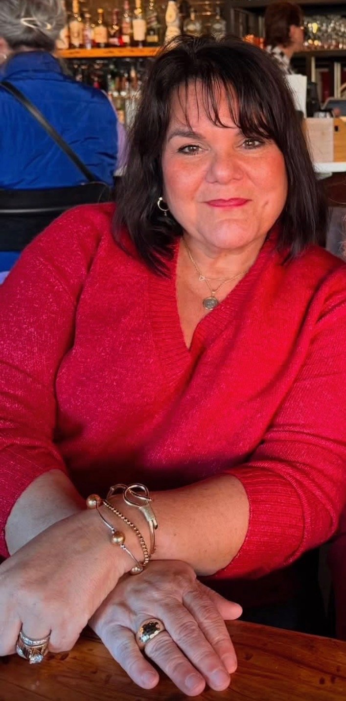 A woman with dark hair wearing a red sweater sitting at a restaurant or bar table with her hands folded. She is smiling slightly and has various jewelry including rings, bracelets, and a necklace. The background shows a bar with bottles and people.