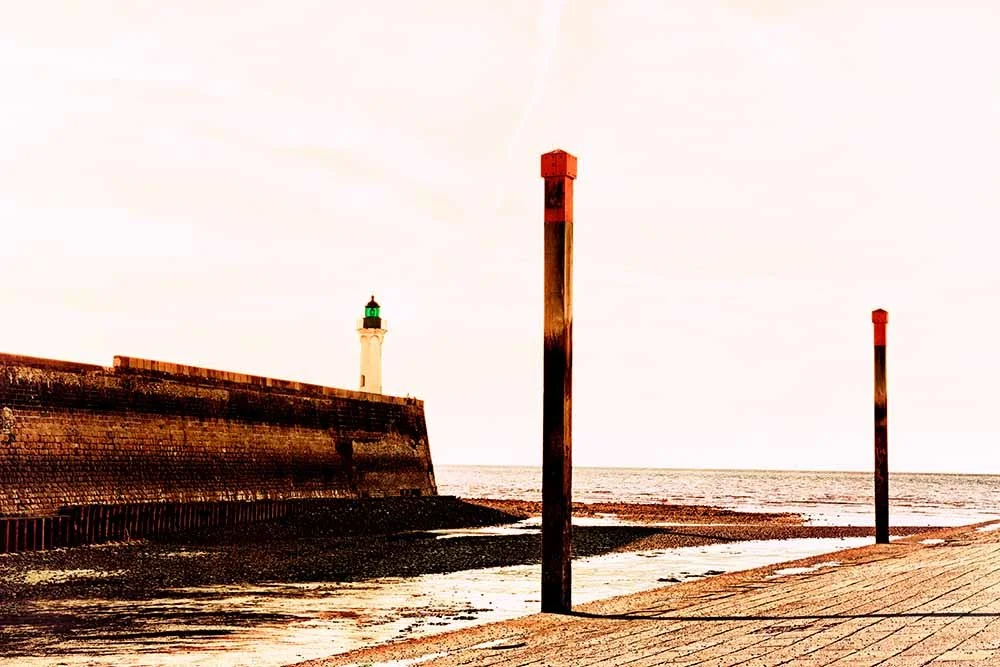 A lighthouse with a green top on a stone pier near the beach, with three wooden posts on the sandy shore and the ocean in the background.
